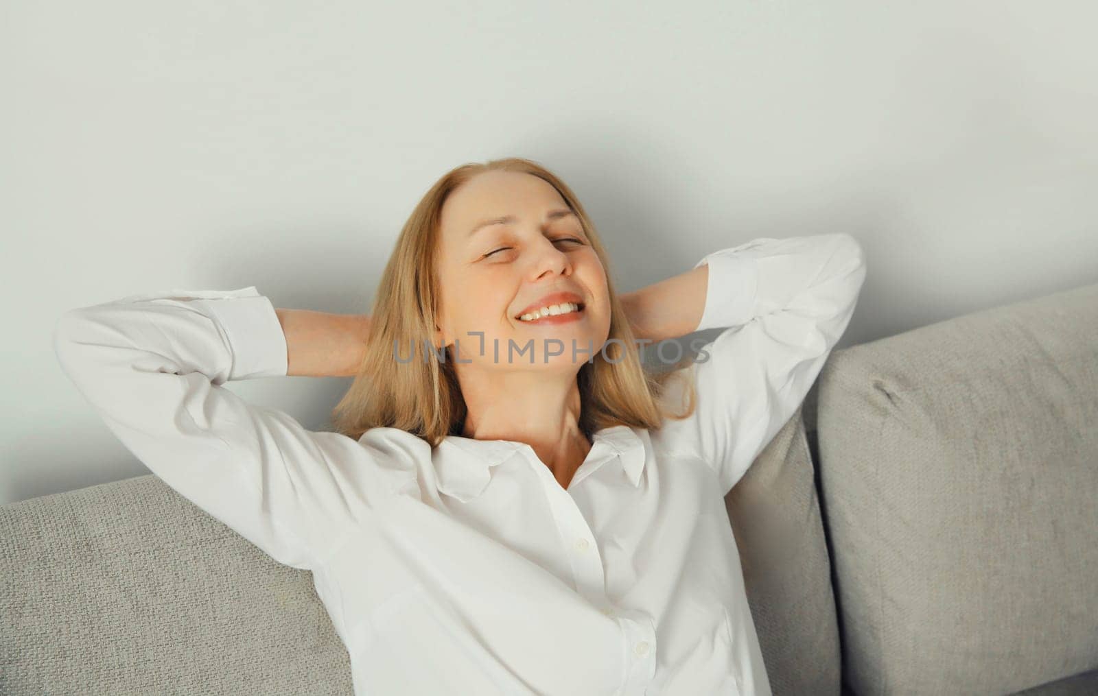 Happy modern relaxed young woman enjoying at home, satisfied girl resting with closed eyes and folded hands behind the head sitting on the couch in living room