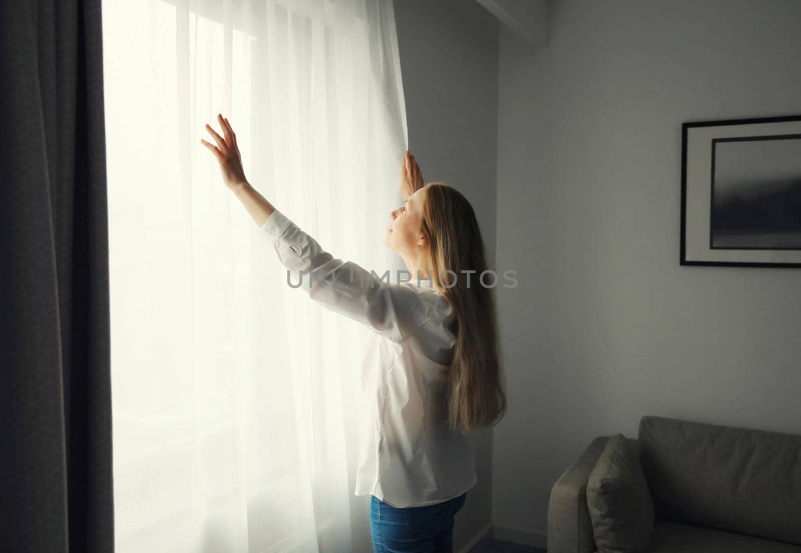 Woman stands by the window, opening curtains at home in morning, household chores, routine or interior decoration after renovation, side view