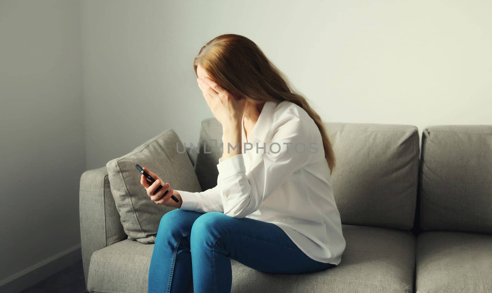 Sad upset crying woman holding phone suffering from stress after reading emotional message sitting on the couch in room at home