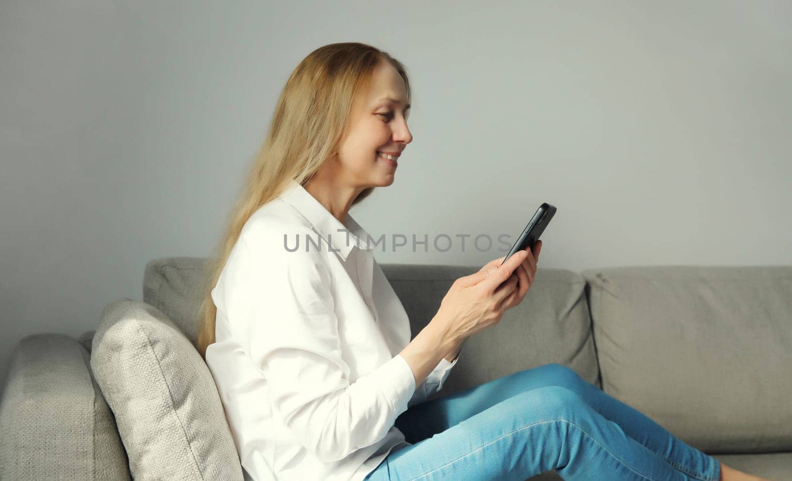 Happy smiling mature woman looking at phone, girl with smartphone sitting on the couch in living room at home