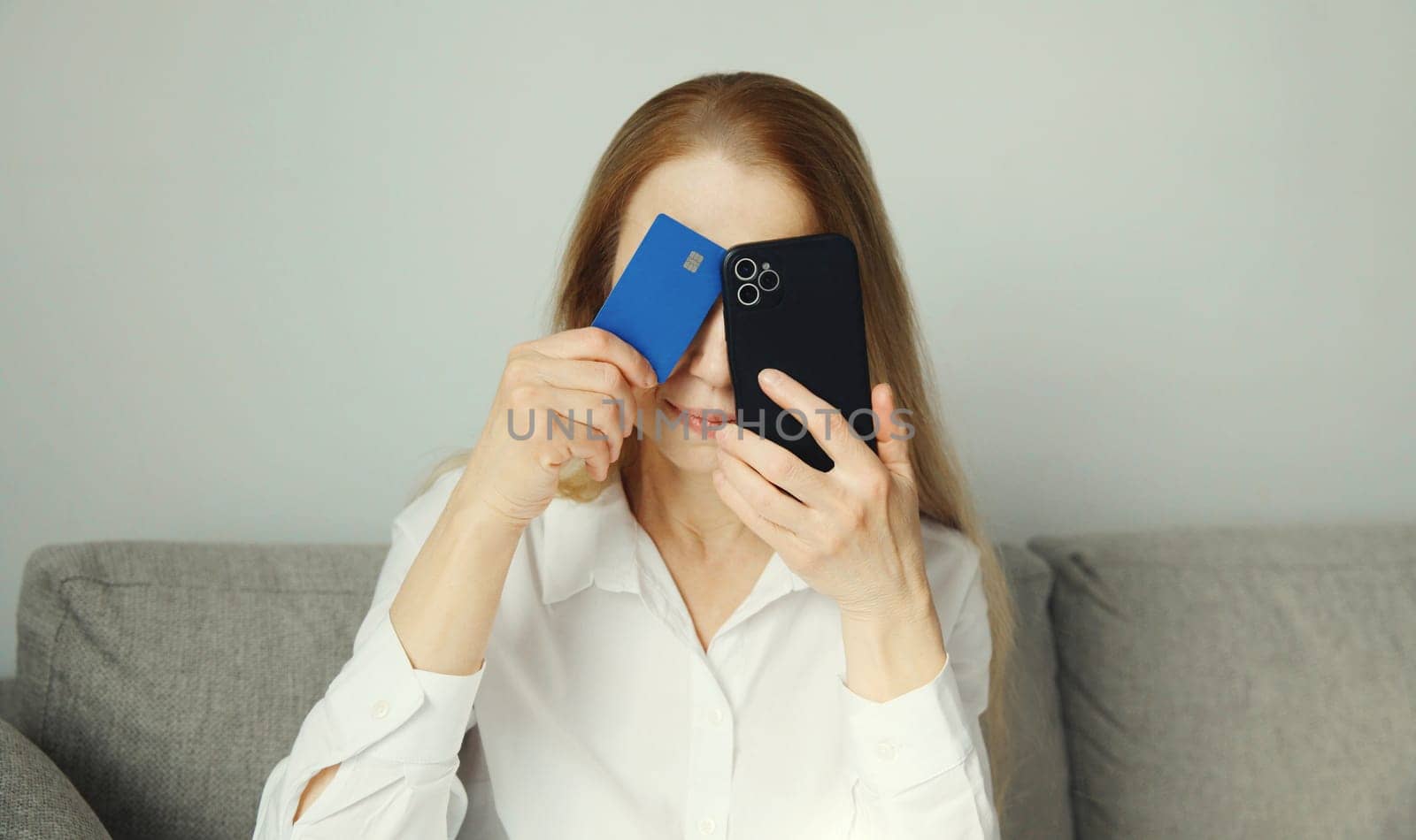 Upset modern young woman holding smartphone and credit card sitting on the couch at home, internet fraud, problems with online payments or blocked bank card