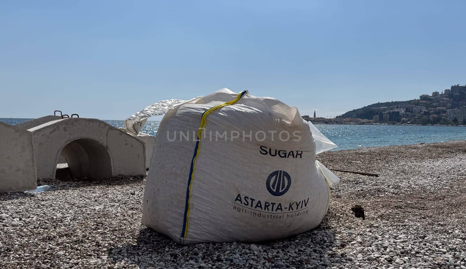 Budva, Montenegro - April 17, 2025: A bag with the inscription SUGAR ASTARTA KYIV, with pebbles and stones inside, stands next to the reinforcing structure of the embankment