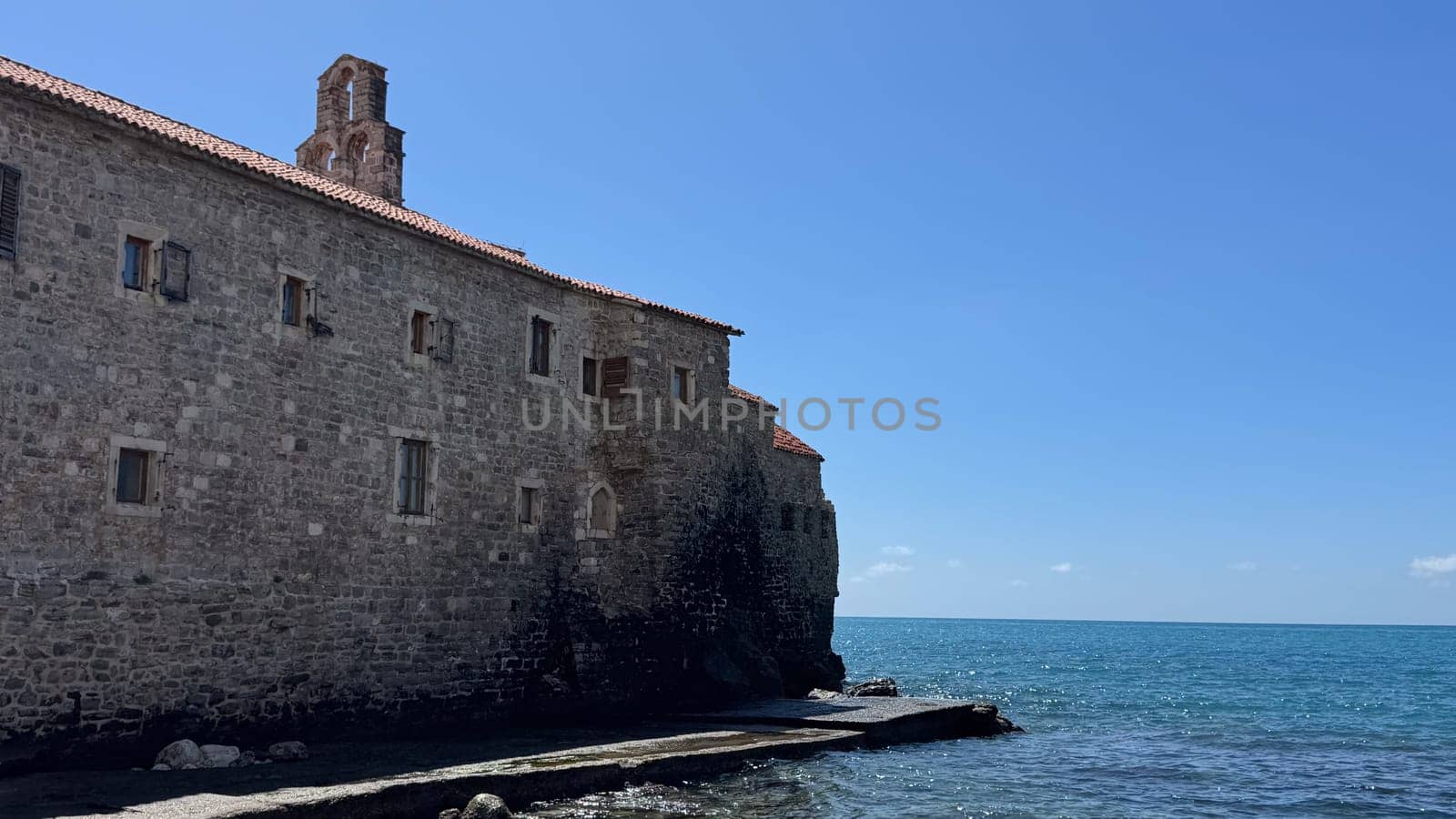 Budva, Montenegro - April 18, 2025: Ancient stone building with a tower The Church of the Virgin Mary above stands on the seashore, surrounded by clear blue water and bright sky