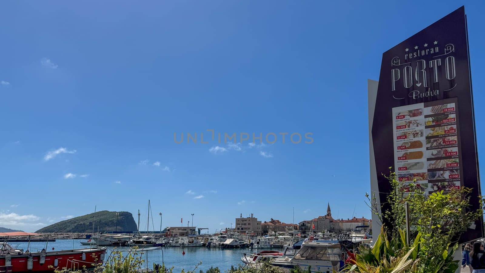 Budva, Montenegro - April 18, 2025: Coastal landscape with a marina filled with boats and a signboard of the PORTO restaurant under a clear sky