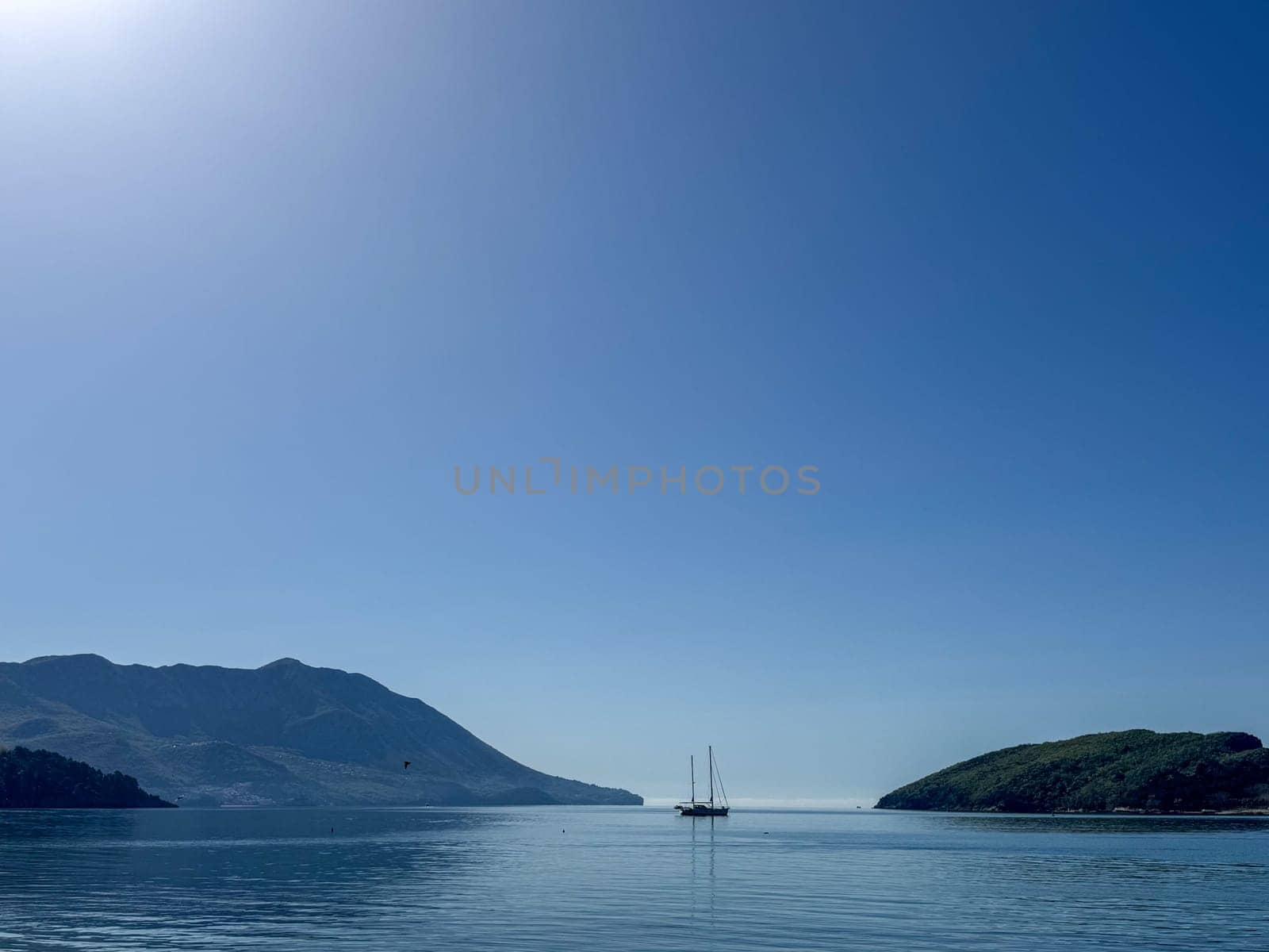 Budva, Montenegro - April 18, 2025: A calm seascape with a sailboat and mountains under a clear blue sky evokes a sense of serenity and peace in nature