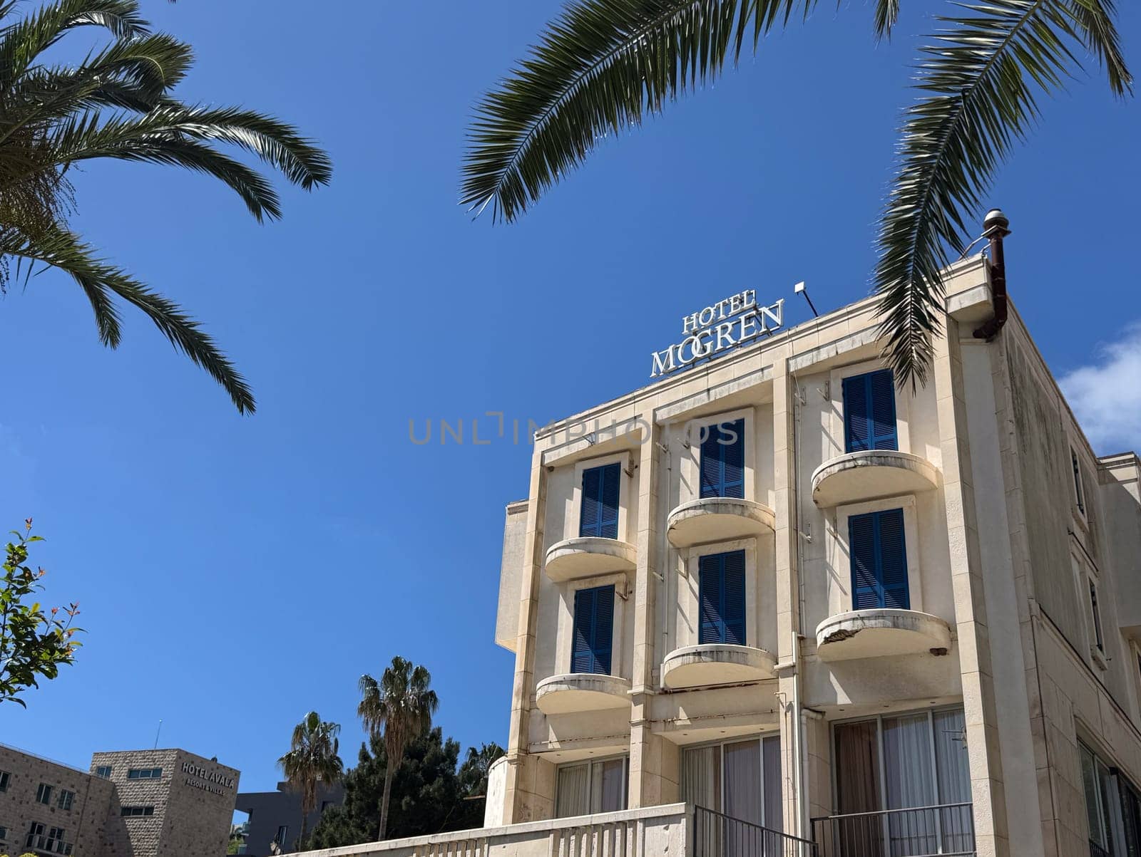 Budva, Montenegro - April 18, 2025: The architectural beauty of the Mogren Hotel against a clear blue sky, surrounded by tropical palm trees, showcasing a serene atmosphere and a welcoming environment