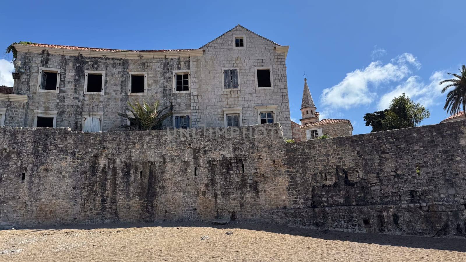 Budva, Montenegro - April 18, 2025: An abandoned historic building located on the seafront, with palm trees and a church tower, embodies history and the passage of time.
