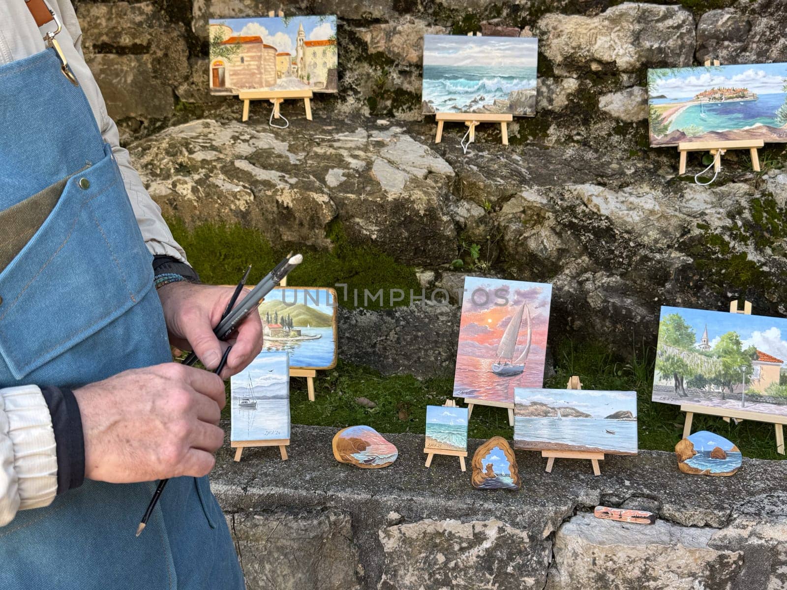 Budva, Montenegro - April 18, 2025: The artist showcases vibrant paintings at an exhibition in a picturesque outdoor setting in the old town. The concept of art evaluation.