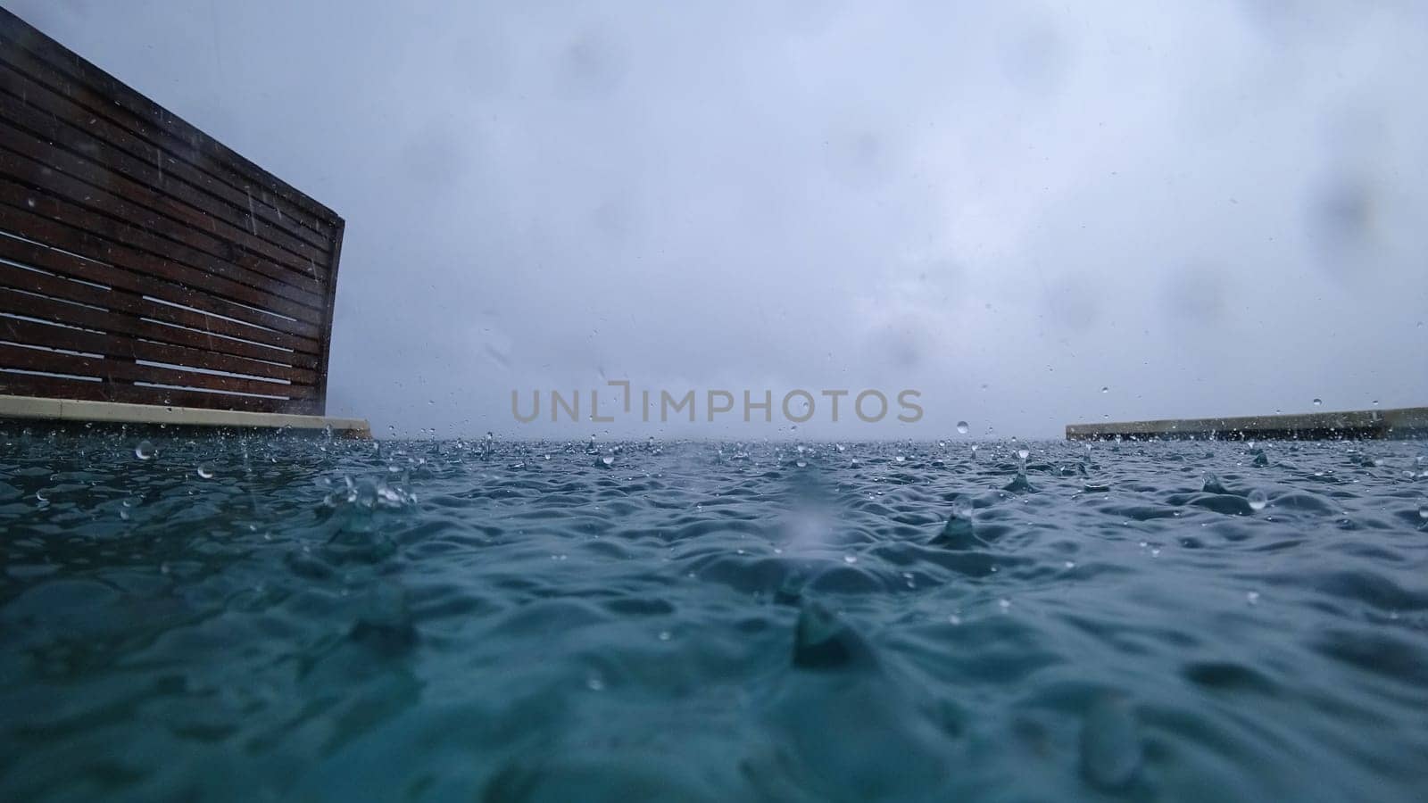 Rain falling on a turquoise swimming pool during a cloudy day creates mesmerizing ripples and splashes on the water's surface, enhancing the tranquil atmosphere of the scene