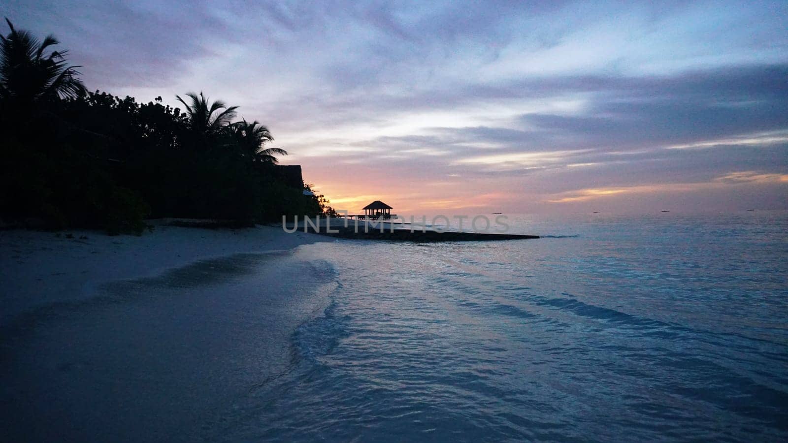 Stunning sunset over a tropical beach in the maldives, with palm trees swaying in the gentle breeze and a gazebo perched over the crystal-clear water, creating a serene and idyllic scene