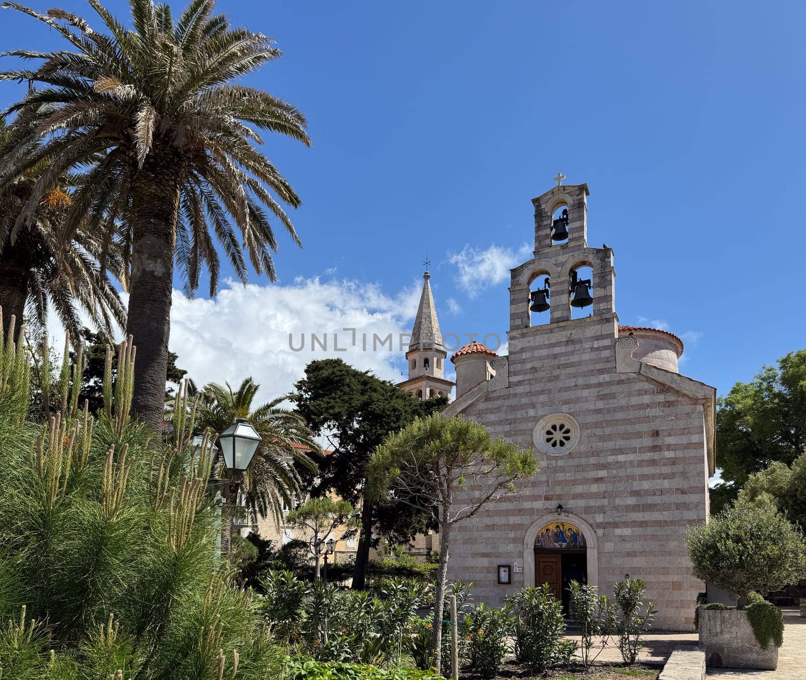 Budva, Montenegro - April 20, 2025, a historic Church of the Holy Trinity in the Old Town, surrounded by palm trees and lush greenery.