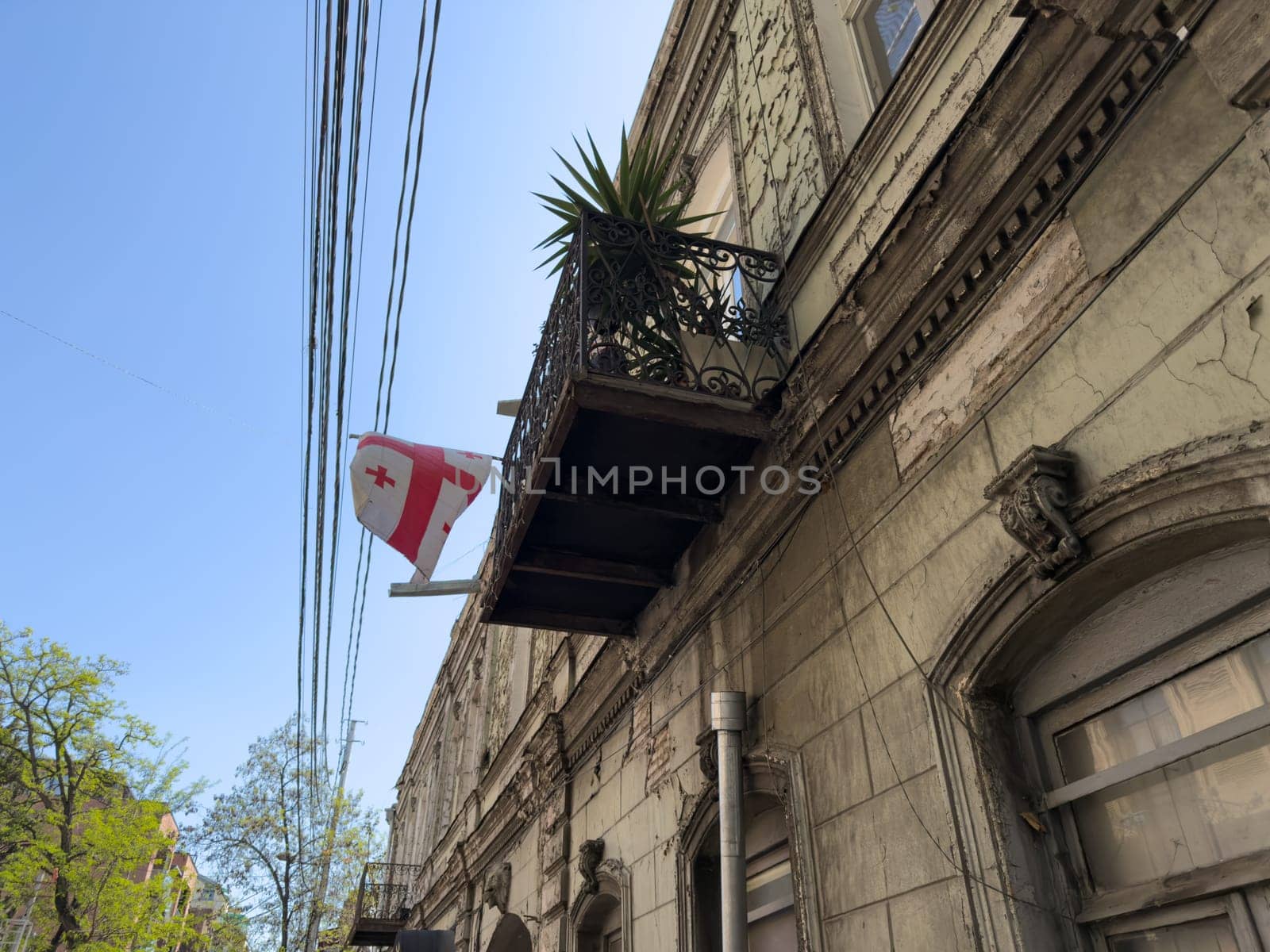 Street view featuring worn down architecture and balcony with potted plant. Power lines stretch across clear sky, capturing real urban life and mood in an older district of Tbilisi, Georgia