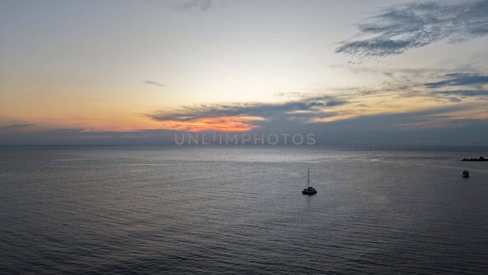 Catamaran sailing peacefully in a calm sea as the sun sets, casting a warm glow on the water and clouds, creating a serene and tranquil atmosphere