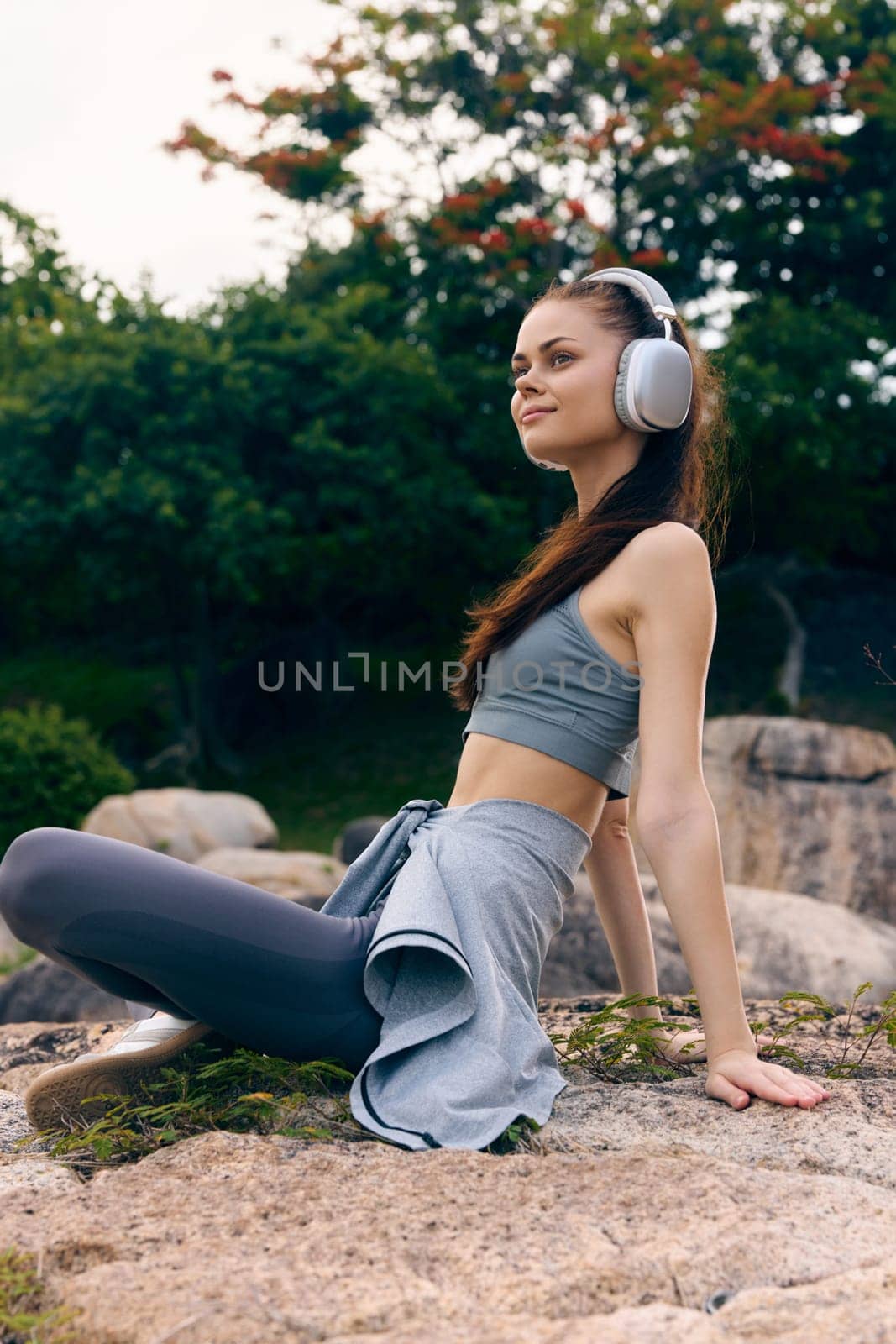 Relaxed young woman in athletic wear with headphones, sitting on a rock in a green outdoor setting, promoting a healthy lifestyle and enjoyment of music