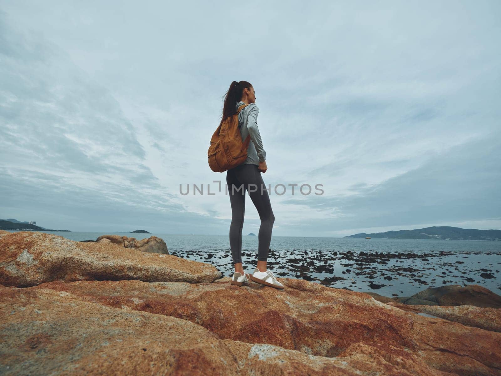 A young woman with a backpack standing on a rocky shore, dressed in athletic wear, embodying exploration and freedom by the ocean, perfect for travel and adventure themes