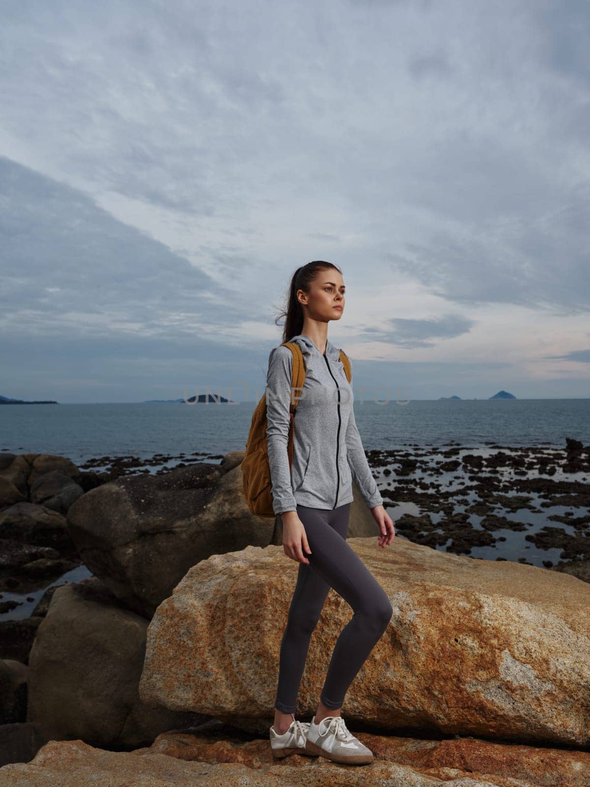 A young woman in athletic clothing stands confidently on rocky seaside terrain, embodying a spirit of adventure and exploration, with a brown backpack and a calm ocean backdrop
