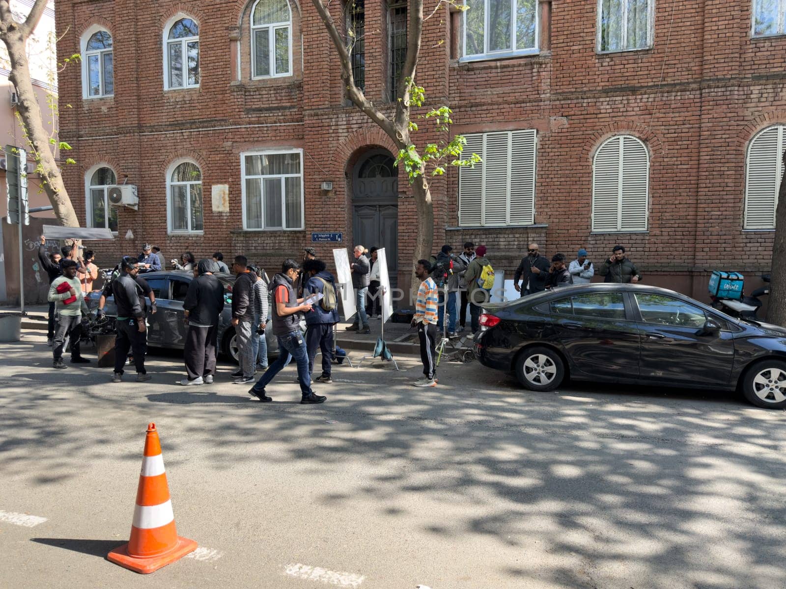 Tbilisi, Georgia, April 11, 2025: Film crew of India ethnicity shoots scene on city street near brick building, with cars, equipment and people preparing for production during sunny day