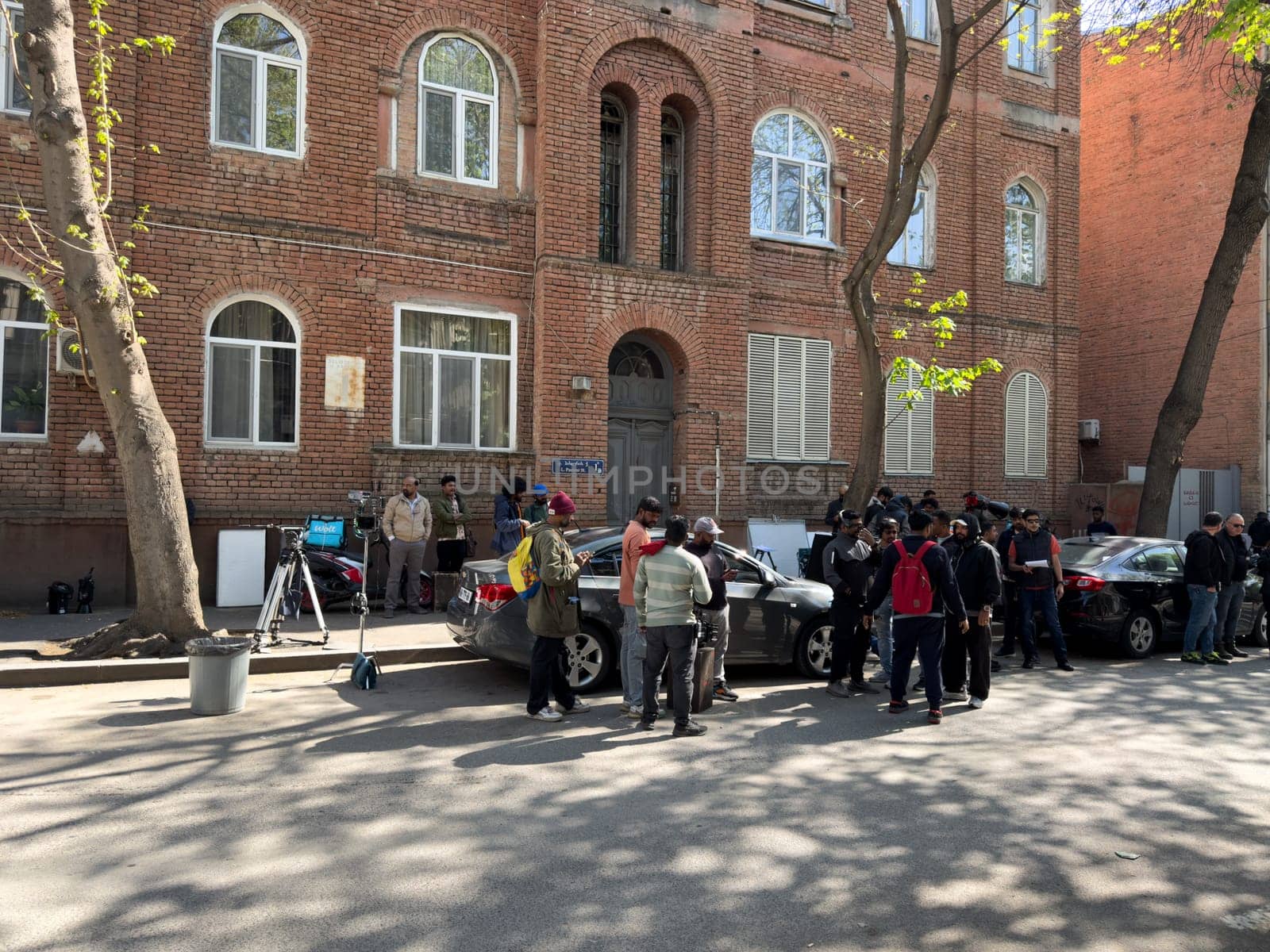 Tbilisi, Georgia, April 11, 2025: Multicultural film team of South Asian ethnicity prepares street shoot near old brick building, using professional equipment, cars and crew coordination on location