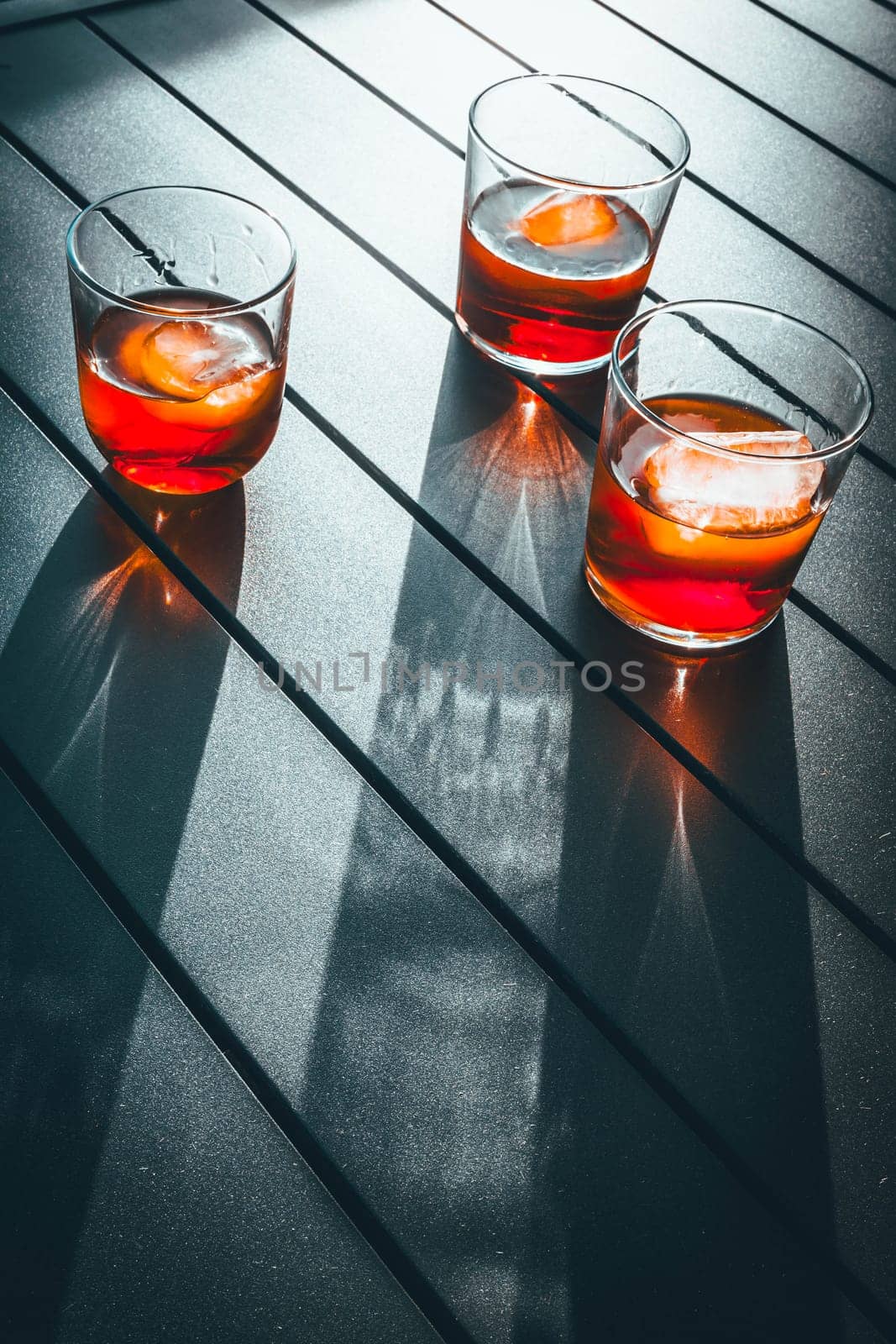 Artistic image of three glasses of red vermouth with ice casting long shadows on a metallic terrace table in bright sunlight. The image captures a Mediterranean lifestyle moment of enjoying a sunny aperitif