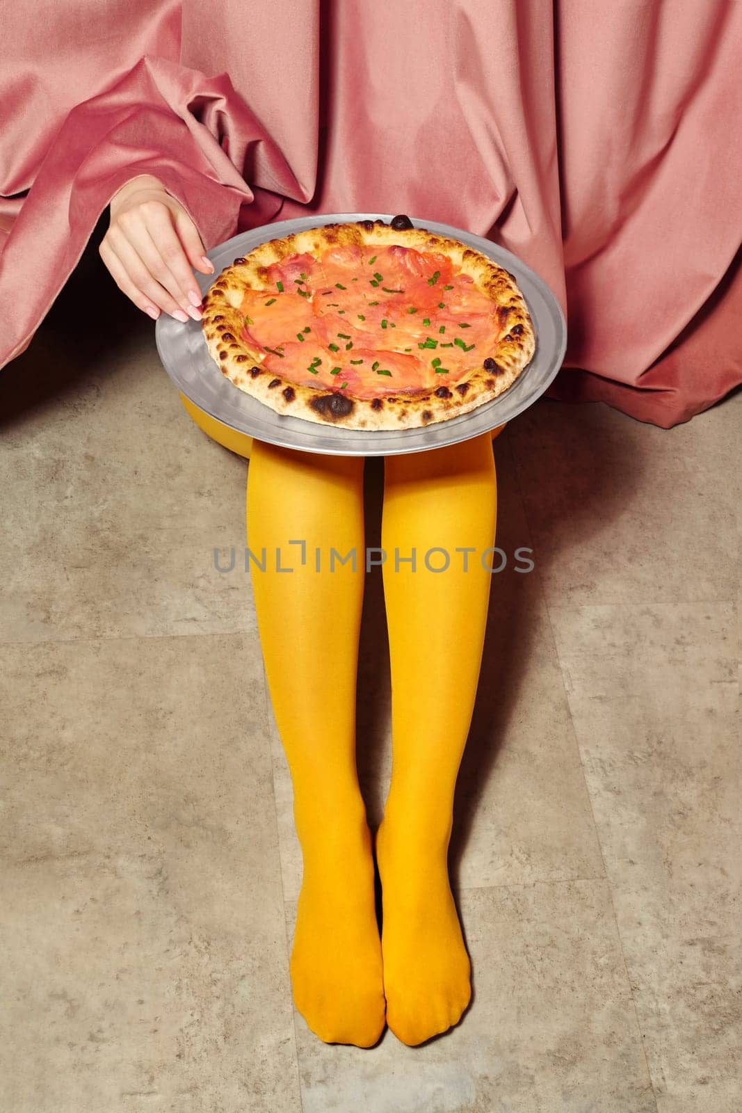 Woman wearing yellow tights presenting a salmon pizza on a tray, hidden behind a pink curtain