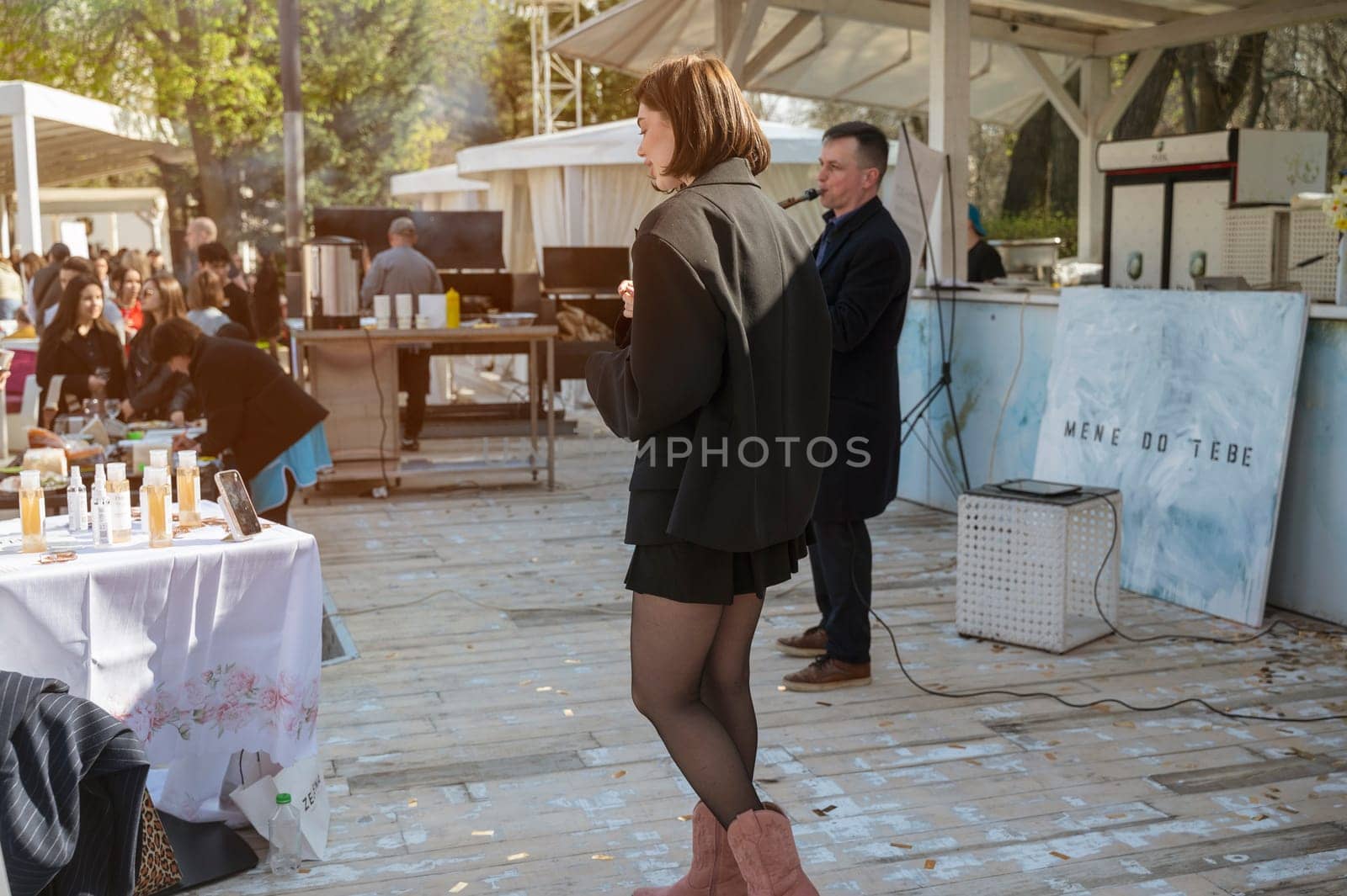 Odessa, Ukraine - April 05, 2025: City food fair. A man in a coat plays the saxophone, a young girl in a jacket and boots dances nearby and films herself on her phone. People enjoy the music