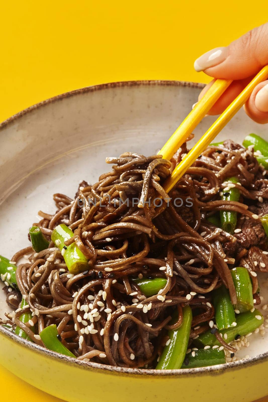 Soba noodles with green beans, beef, and sesame seeds are picked up with yellow chopsticks from a bowl on a vibrant yellow background