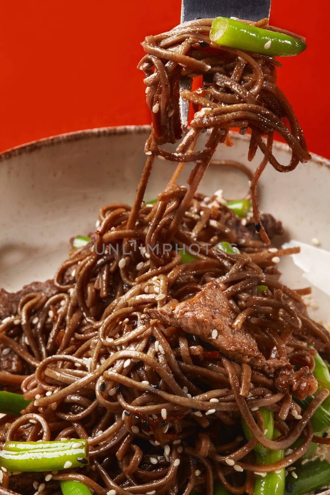 Soba noodles with beef and green beans sprinkled with sesame seeds are lifted by a fork from a bowl on a red background