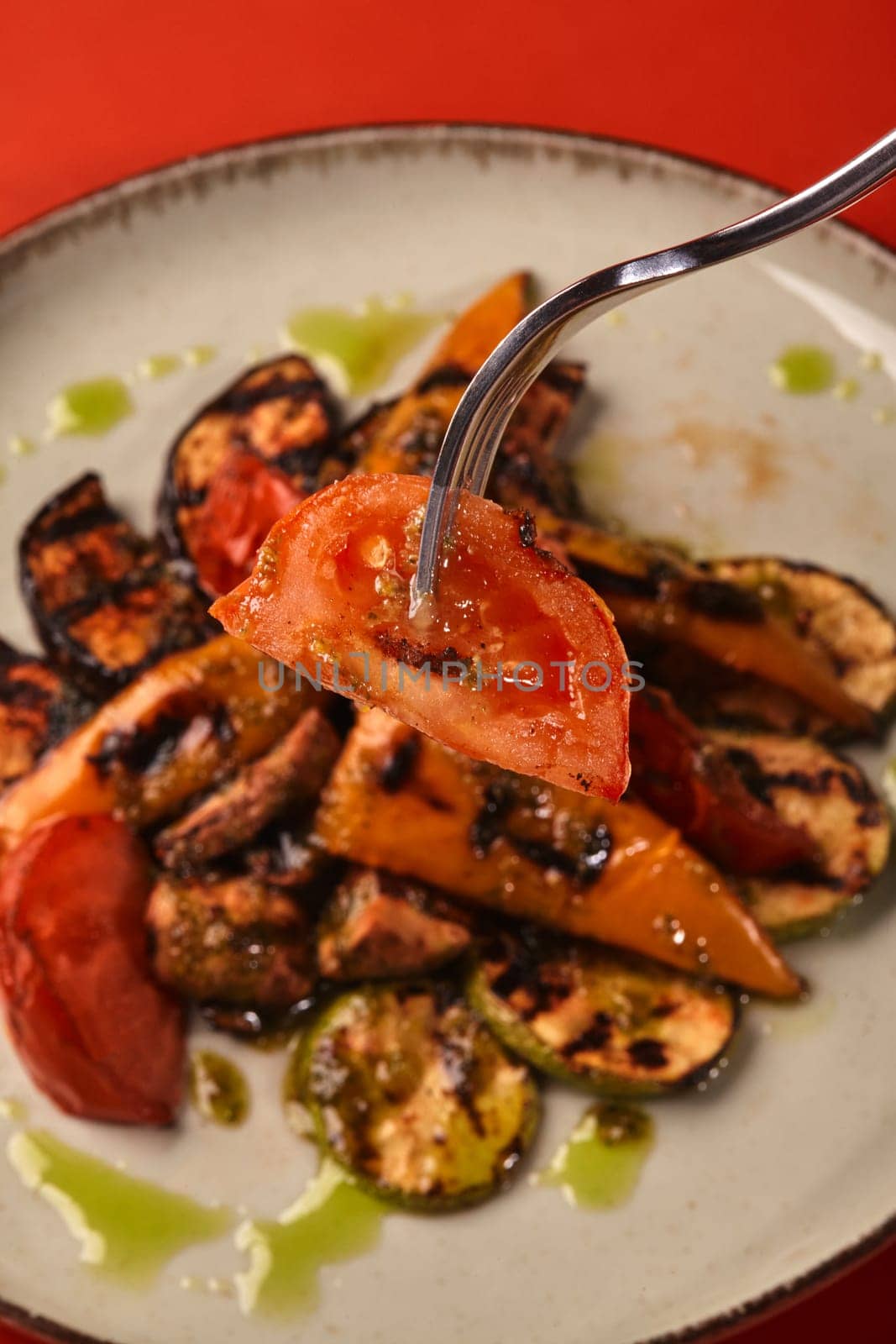 Grilled vegetables with pesto sauce, including tomatoes, zucchini, eggplant, bell peppers, and mushrooms, presented on a plate, with a fork lifting a tomato slice