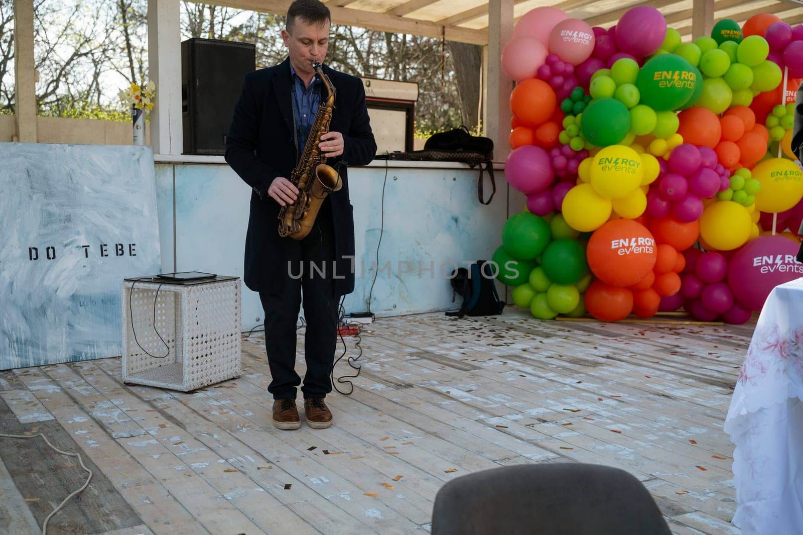 Odessa, Ukraine - April 05, 2025: City food fair. A man in a coat plays the saxophone, on a makeshift stage made of white wood and with colorful balloons