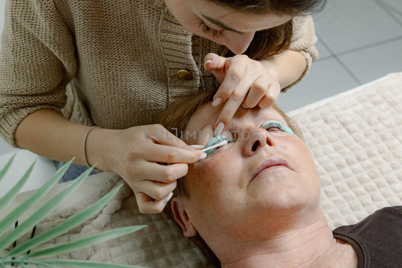 One Caucasian young beautiful brunette cosmetologist wipes excess adhesive with a cotton stick on the eyelashes of the right eye with a sponge stick to an elderly female client who is lying on the cosmetology table in a home beauty salon, side view very close-up.