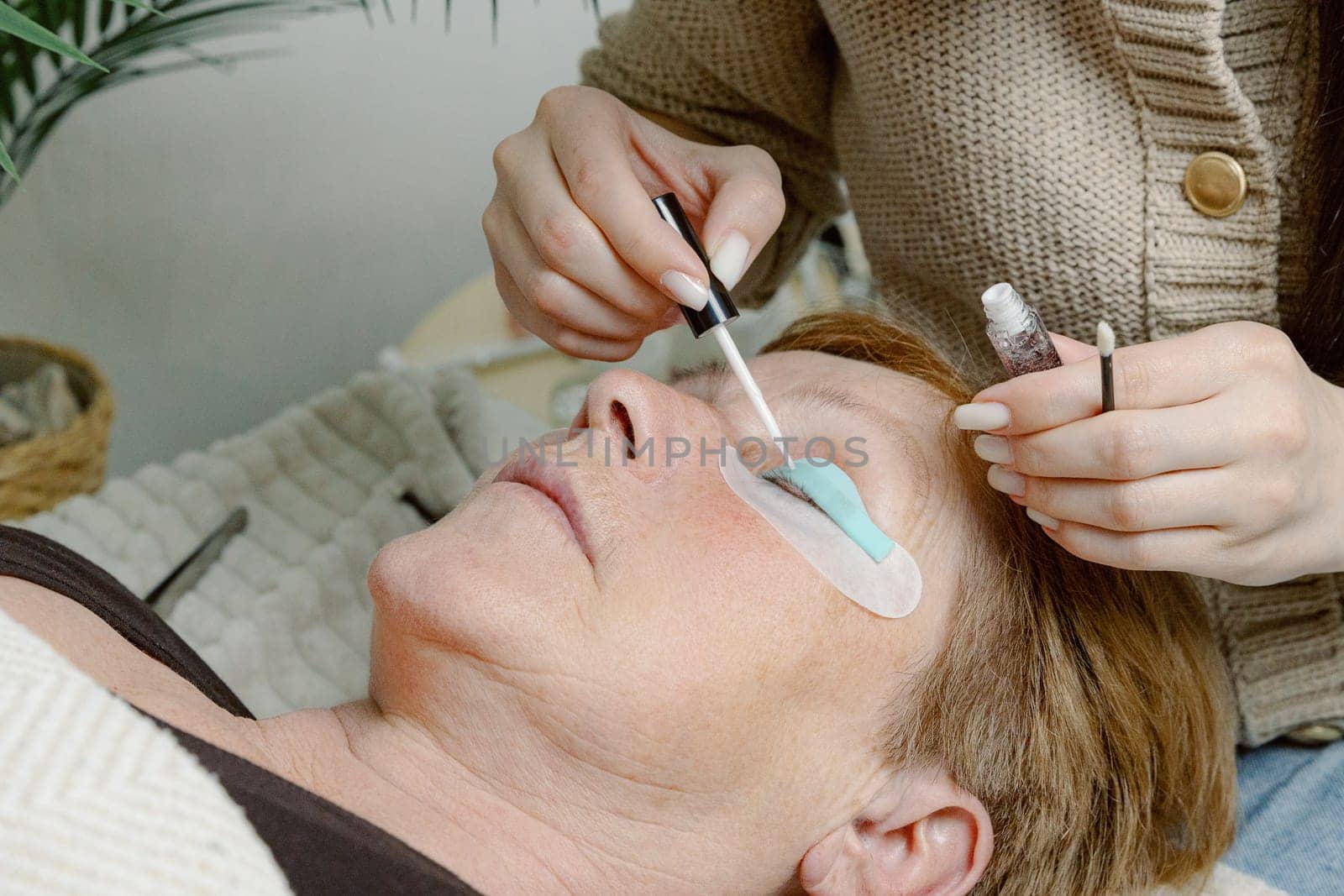 One Caucasian young unrecognizable girl cosmetologist applies glue to a silicone stand for the eyelashes of the left eye using a brush to an elderly female client who is lying on a cosmetology bed in a home beauty salon, side view close-up.