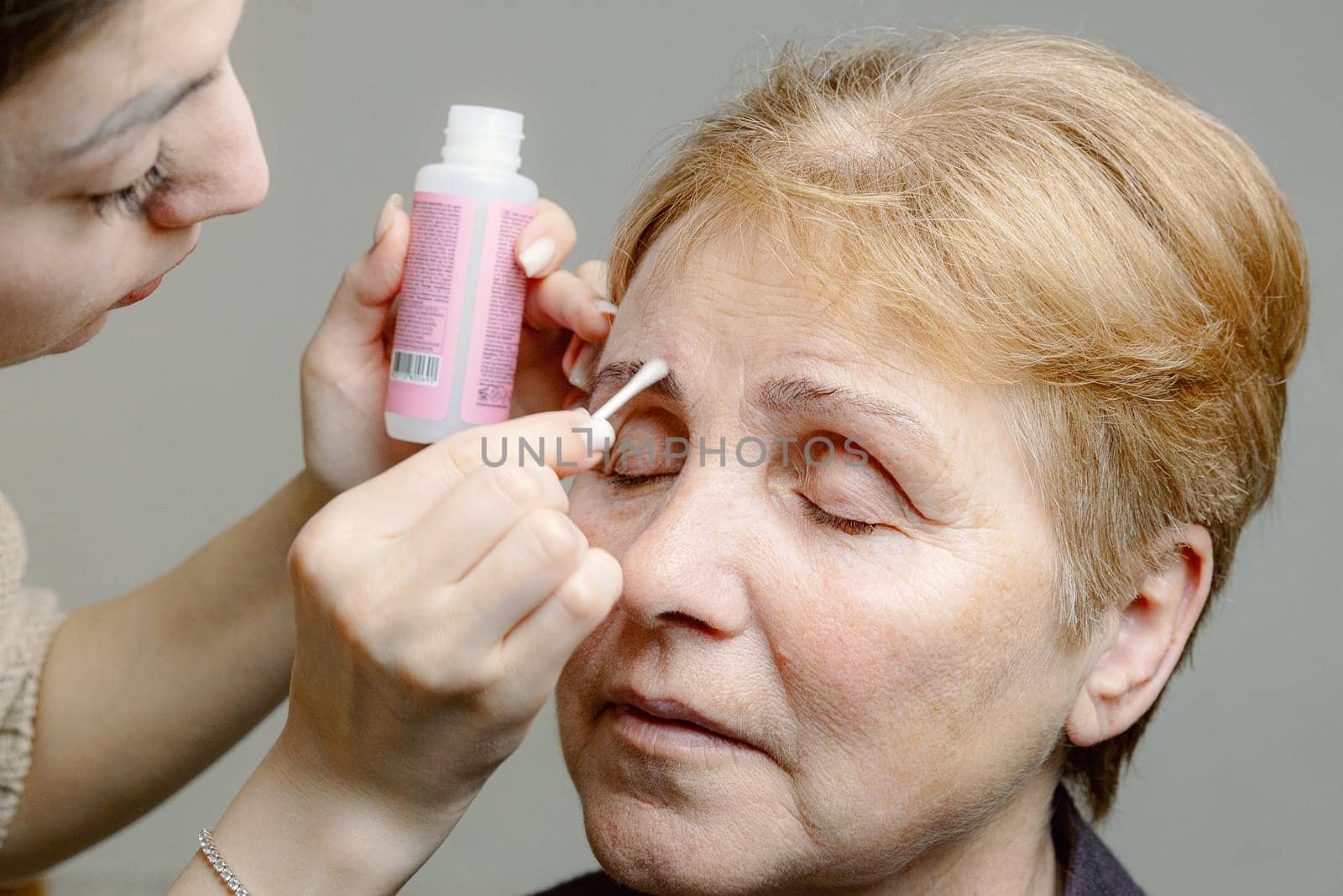 One young Caucasian young beautiful girl cosmetologist carefully removes with a cotton stick the remaining paint from the right eyebrow of an elderly woman with closed eyes, looking to the side using a cleanser, in a home beauty salon, close-up side view.