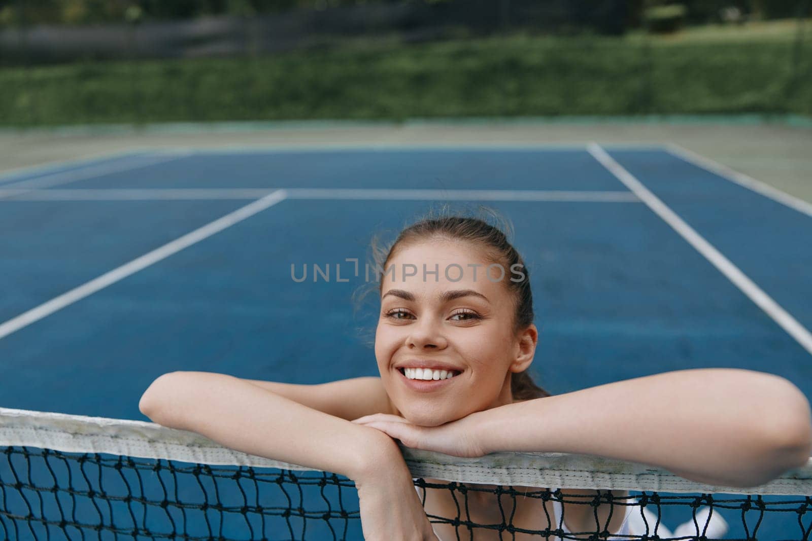 Happy young female tennis player leaning on the net, radiating energy and passion for the sport, with a vivid blue tennis court background ideal for health and fitness themes