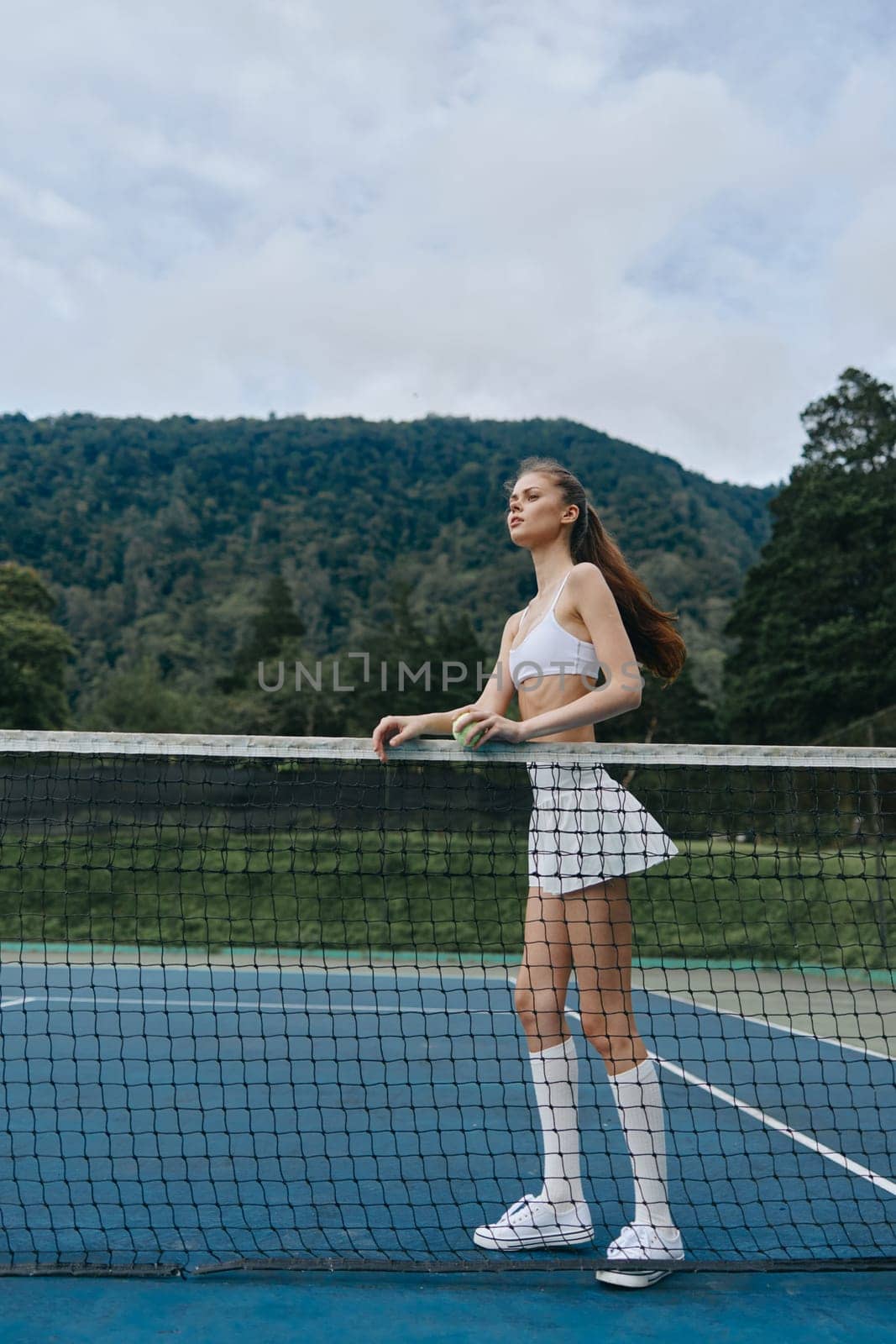 Energetic young female tennis player in a stylish white outfit exudes confidence on the court, set against a lush green mountainous background, ideal for sports promotion