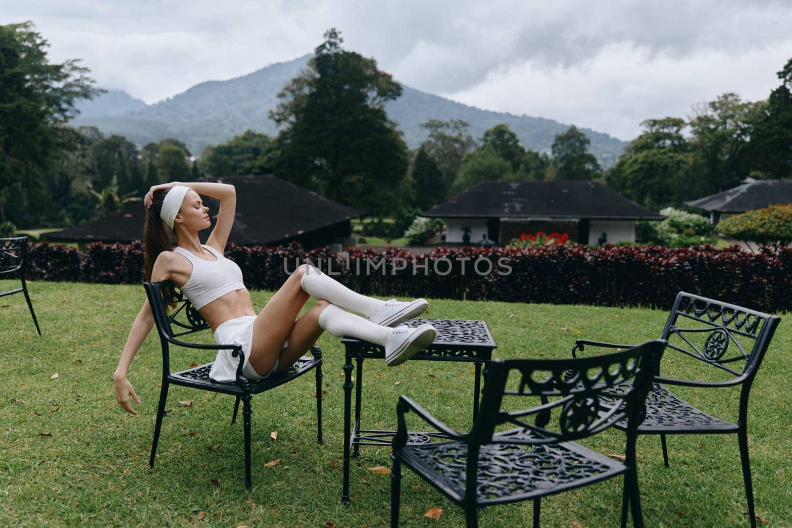 A fashion forward model dressed in a sporty ensemble, exuding relaxation and confidence while lounging on a chair amidst lush green hills and cloudy skies