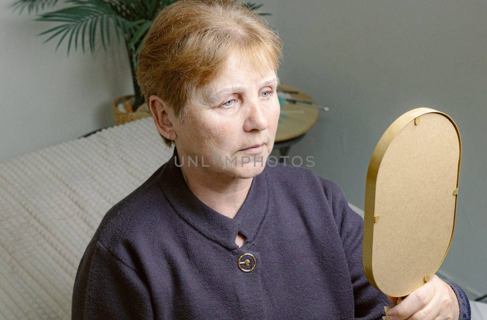 One young Caucasian elderly woman looks in the mirror, carefully examining the drawn white outline on her eyebrows before starting coloring while sitting in a home beauty salon on a cosmetology bed, top side view close-up.