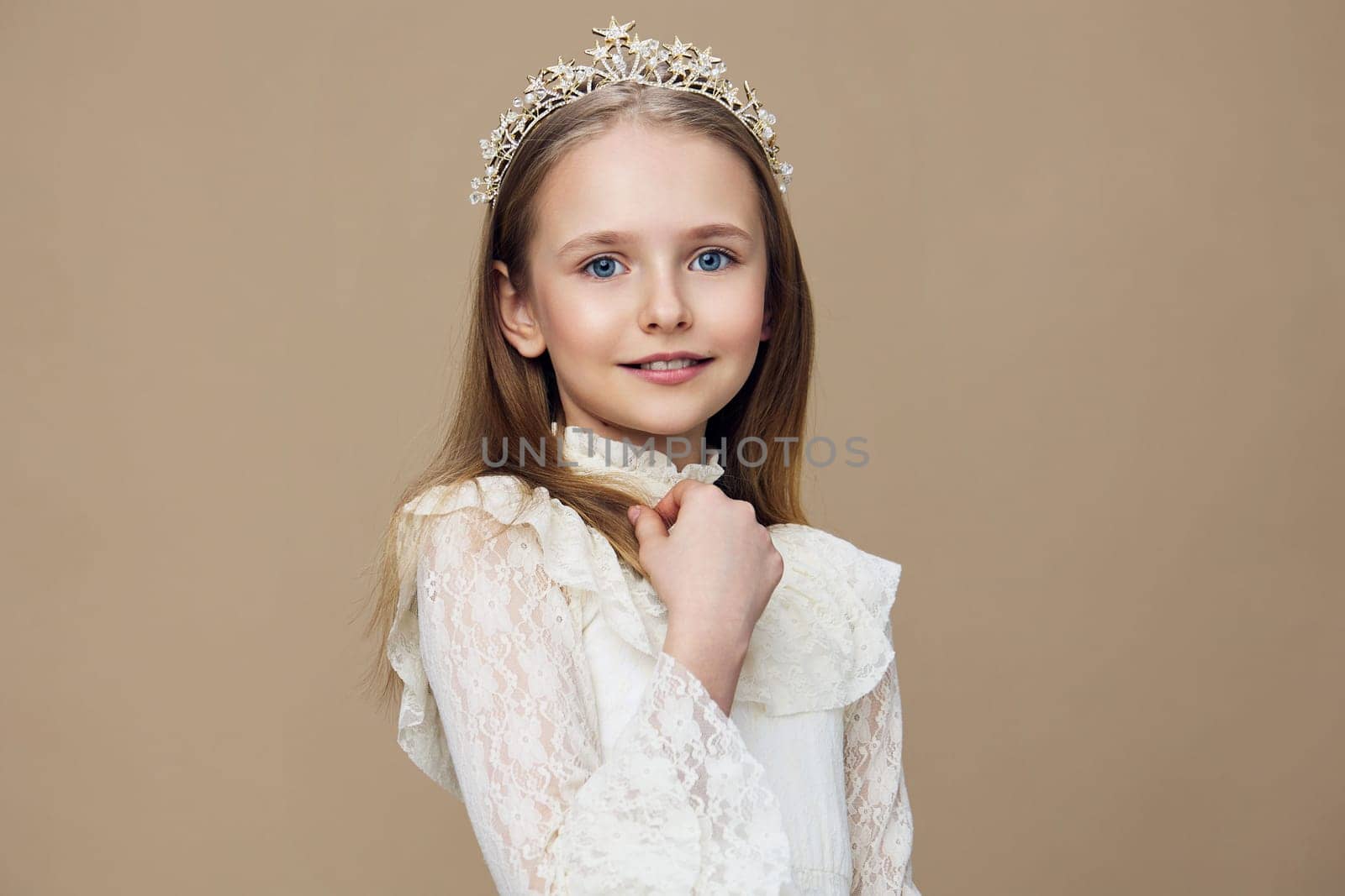 This portrait features a lovely young girl in a delicate lace dress and a crown, exuding a sense of joy and innocence The warm beige background enhances the overall softness and charm, ideal for children s portrait photography