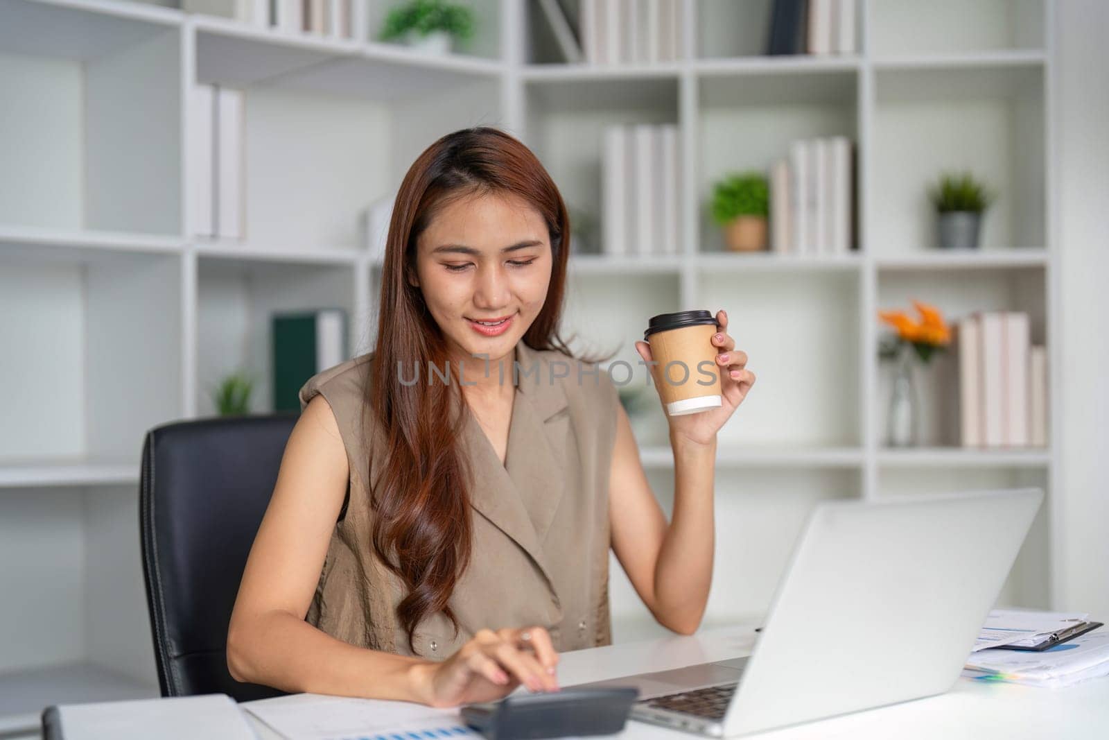 A focused young professional sits in her modern office workspace, sipping coffee and calculating important figures on her calculator, showcasing a productive work atmosphere.
