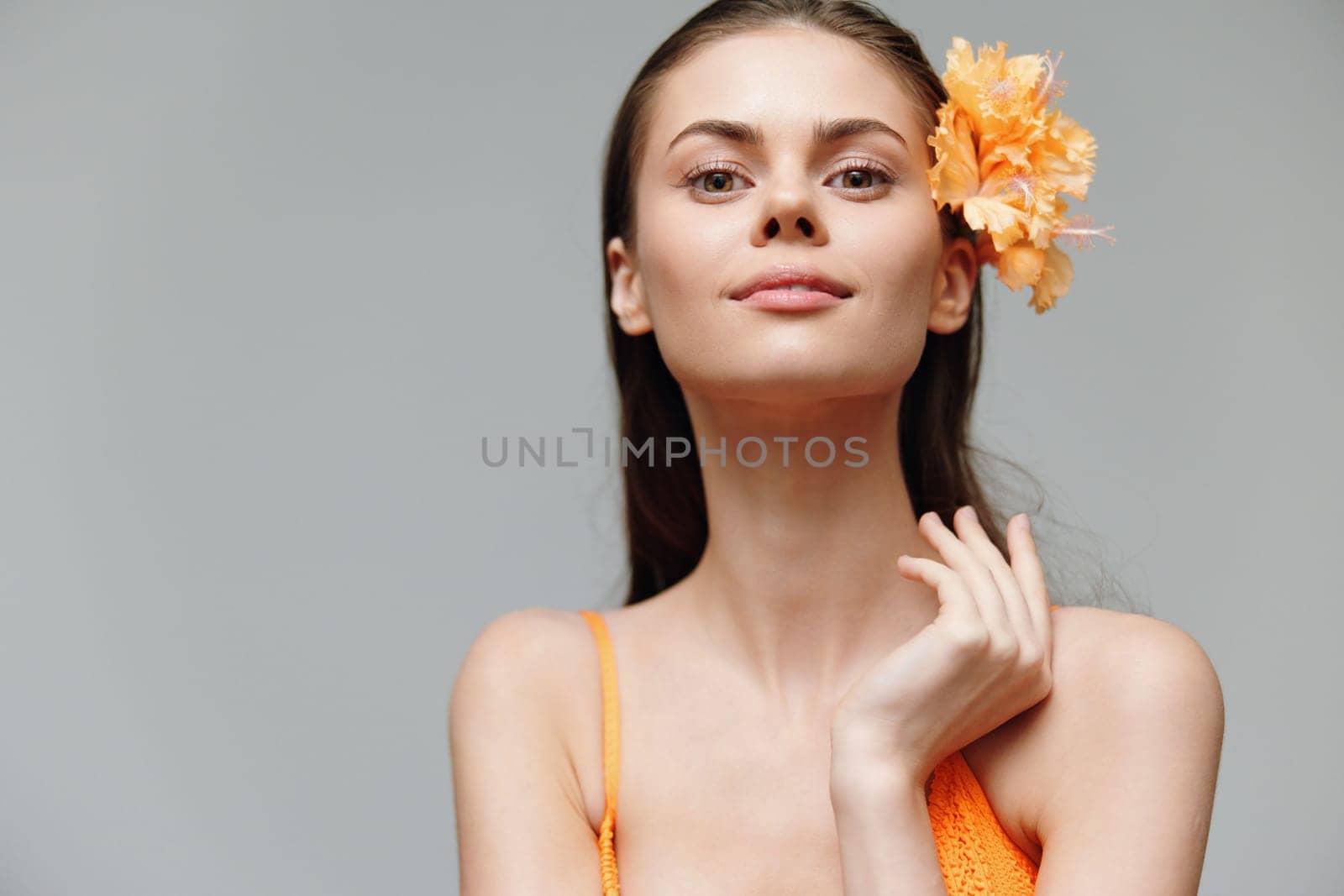 A stunning young woman poses gracefully with a bright orange outfit and a flower in her hair, showcasing beauty and confidence against a soft gray background, ideal for fashion and editorial use.