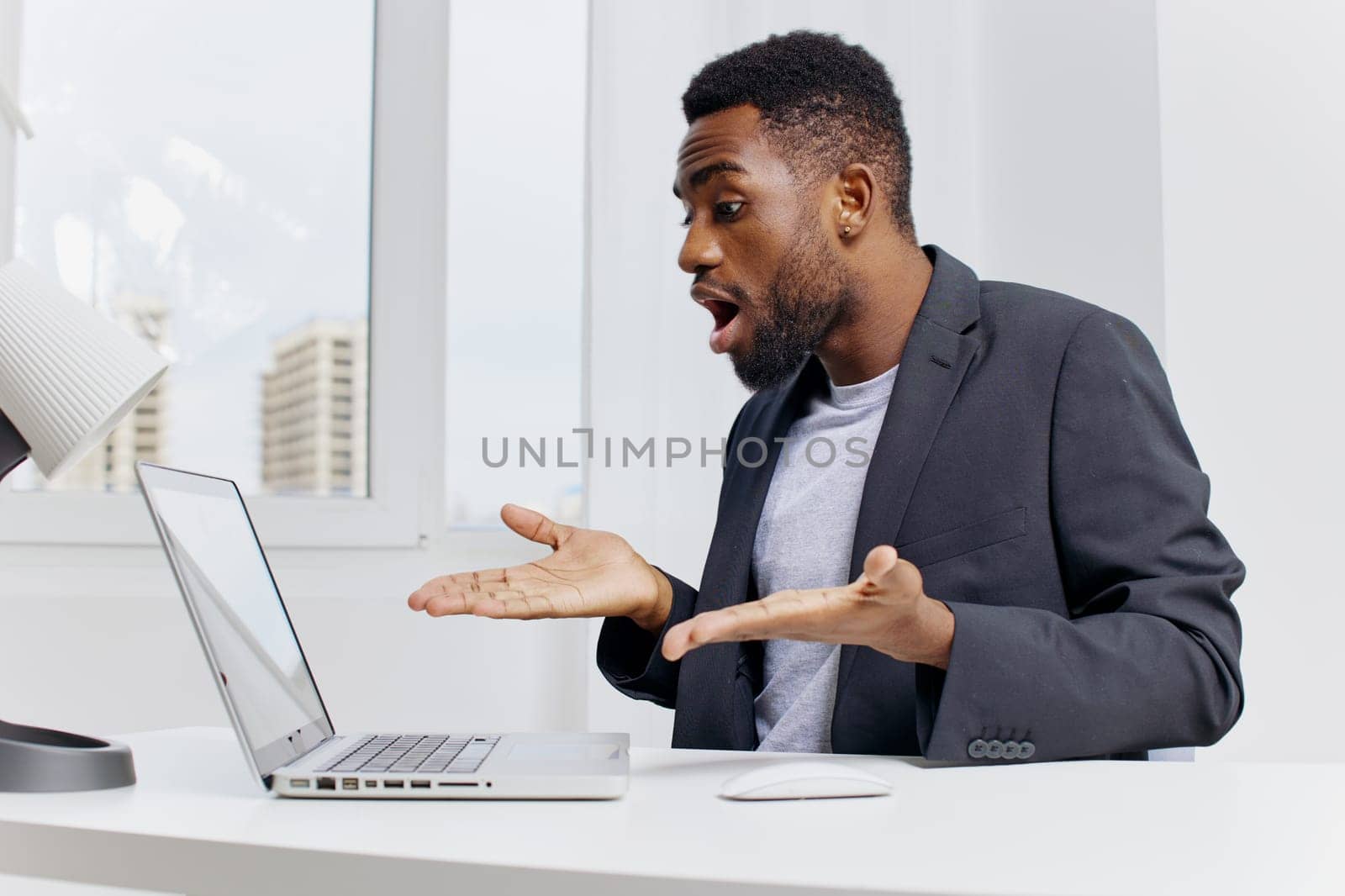 Stressed businessman in formal attire holding head in frustration while working on laptop at cluttered office desk.
