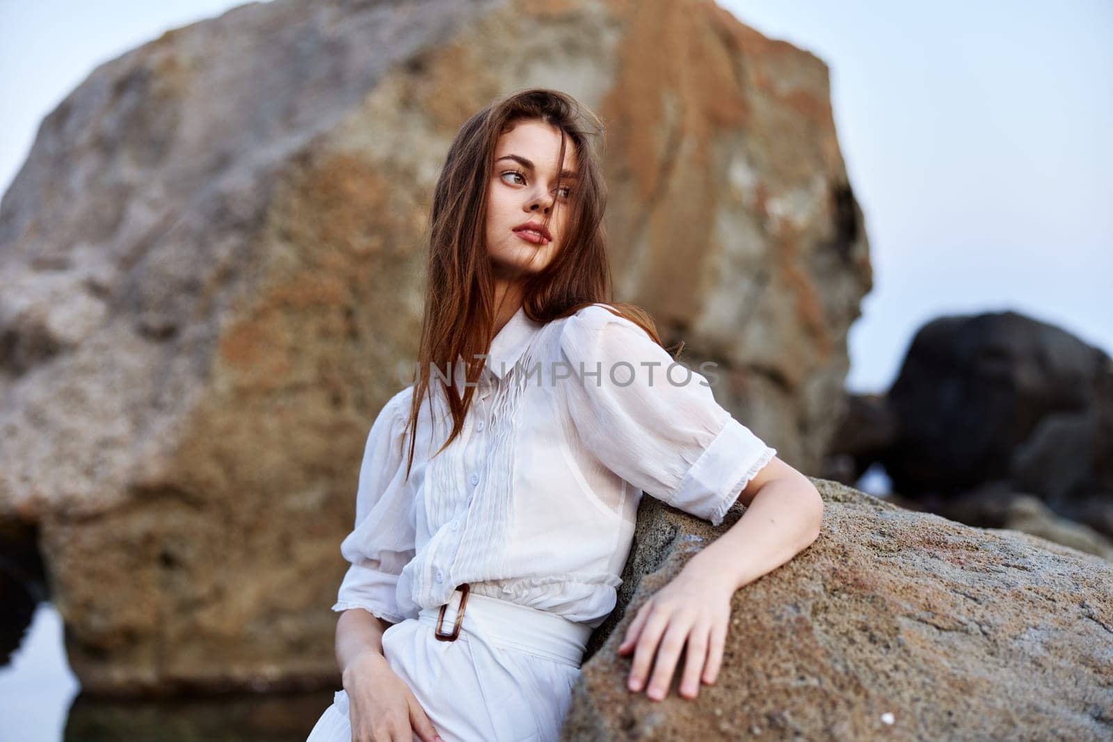 Serene woman in flowing white dress strikes confident pose on ocean rock