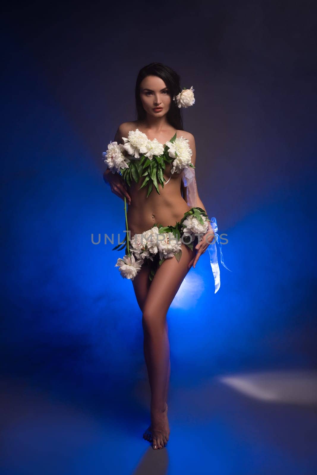 Gorgeous young self assured female model with long dark hair and fresh white flowers covering naked body standing in dark studio - smoke under neon light