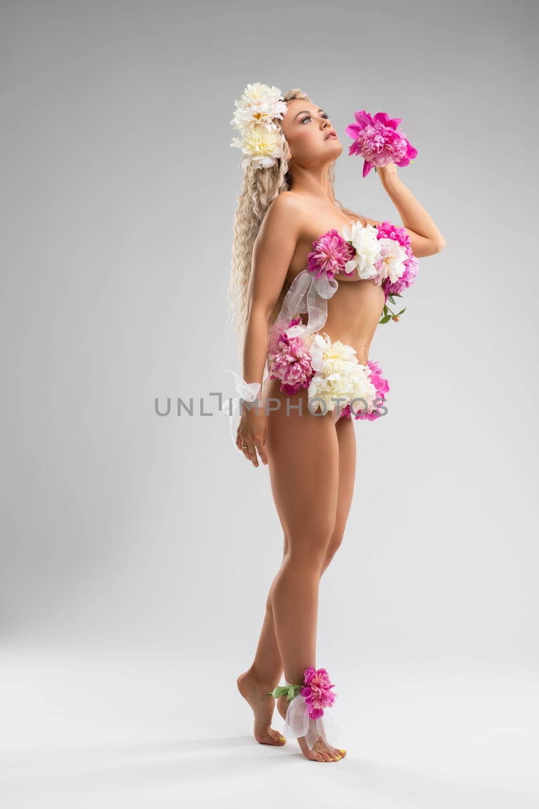 Side view of sensual young female in white lace lingerie and floral wreath looking at camera against gray background