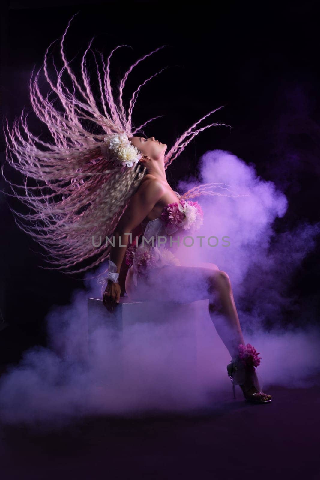 Side view of gorgeous young female dancer in floral bra shaking long blond curly hair while sitting on cube on dark stage with purple fume