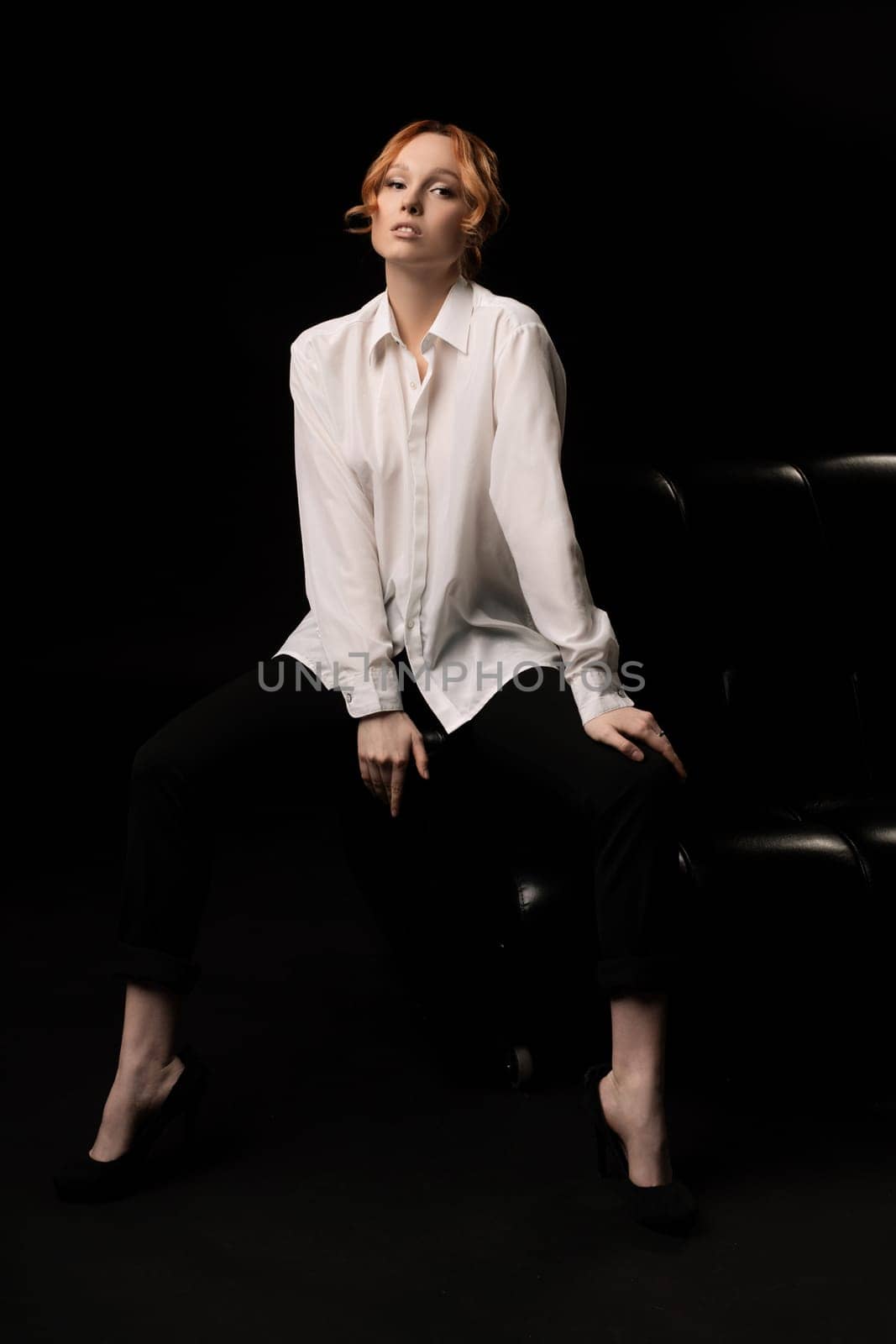 Full body of confident serious young female manager with ginger hair in elegant transparent white shirt sitting on comfortable leather sofa