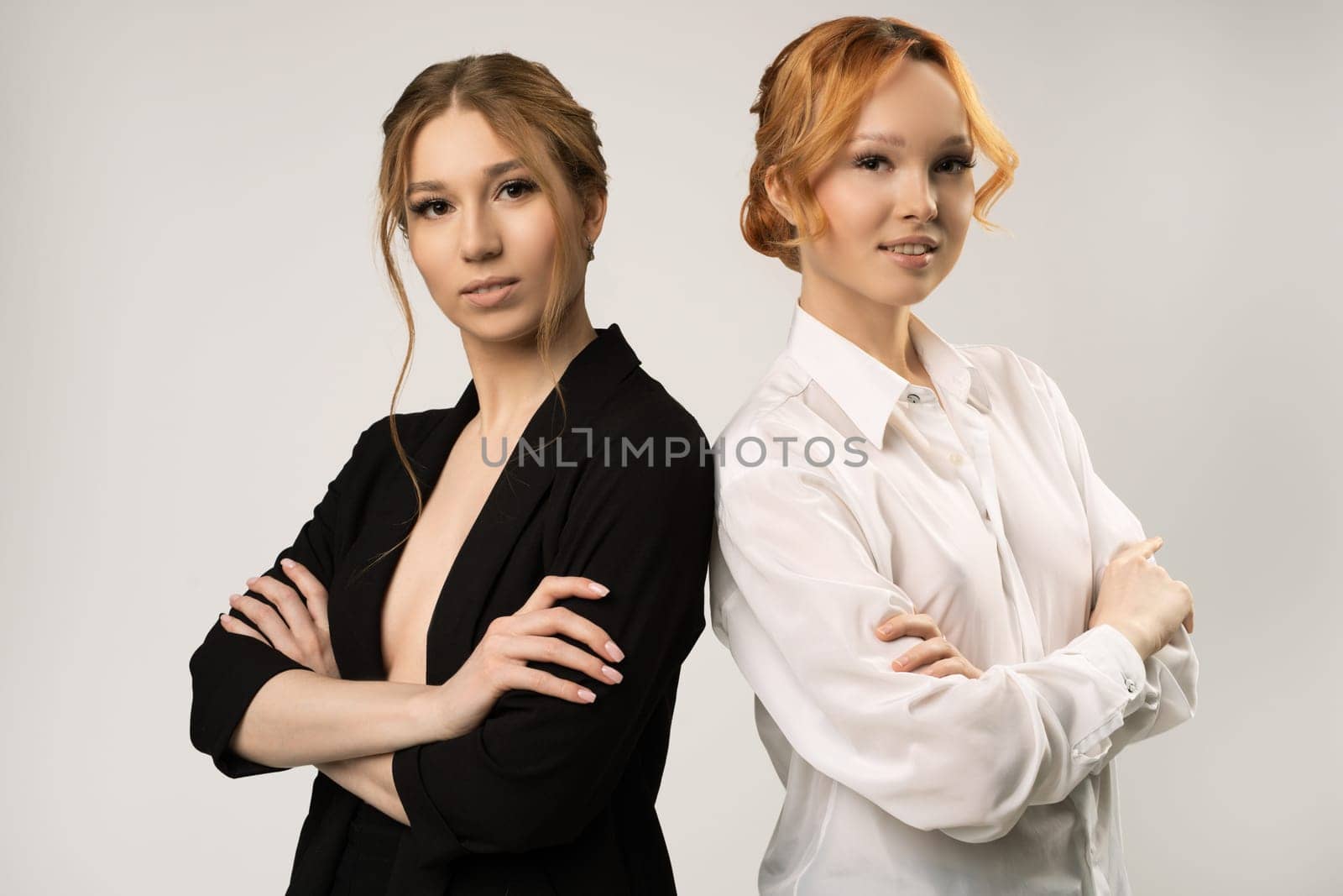 Positive confident young female colleagues in elegant outfits standing back to back with crossed arms and looking at camera against gray background