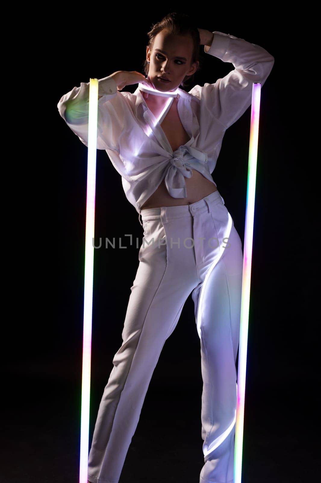 Confident young lgbt male model in white shirt and pants with makeup touching face and looking at camera while standing under neon lights
