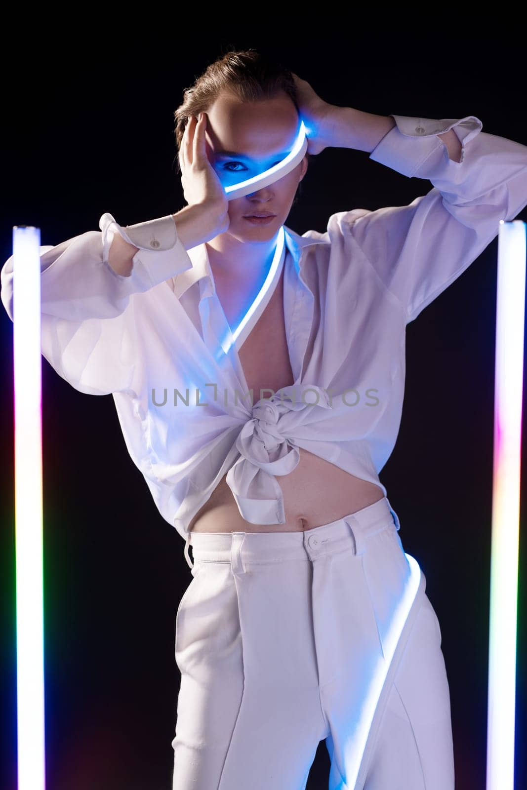 Confident young androgynous male model in white shirt and pants with makeup touching face and looking at camera - black studio near LED sticks