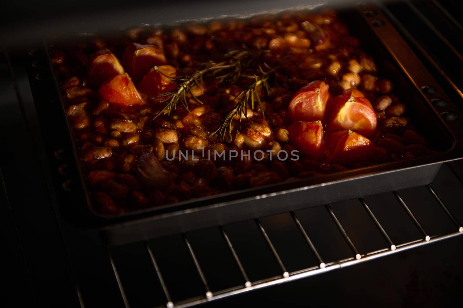 A baking dish filled with beans, tomatoes, and herbs is baking in an oven under warm light. The rustic preparation creates an inviting atmosphere, highlighting the freshness and simple cuisine.