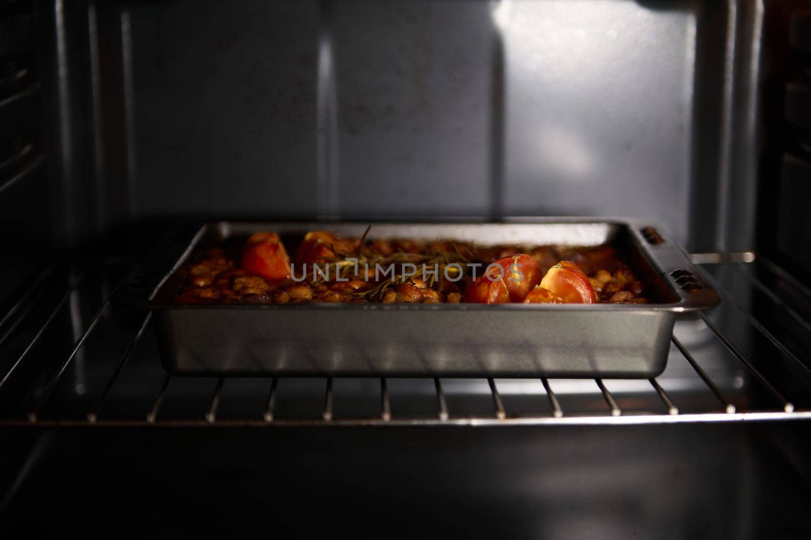 A close-up view of a roasted dish featuring beans and tomatoes, garnished with fresh rosemary inside an oven. The image captures the golden hues and rustic ambiance of home-cooked meals.